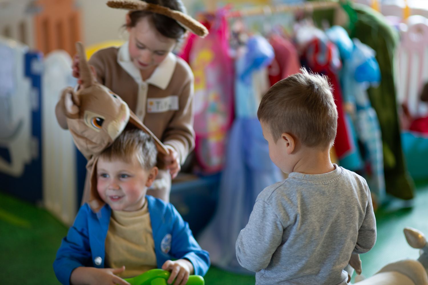 Three children in a play area, one putting a bunny hat on another. One child looks on. Colorful costumes hang in background.