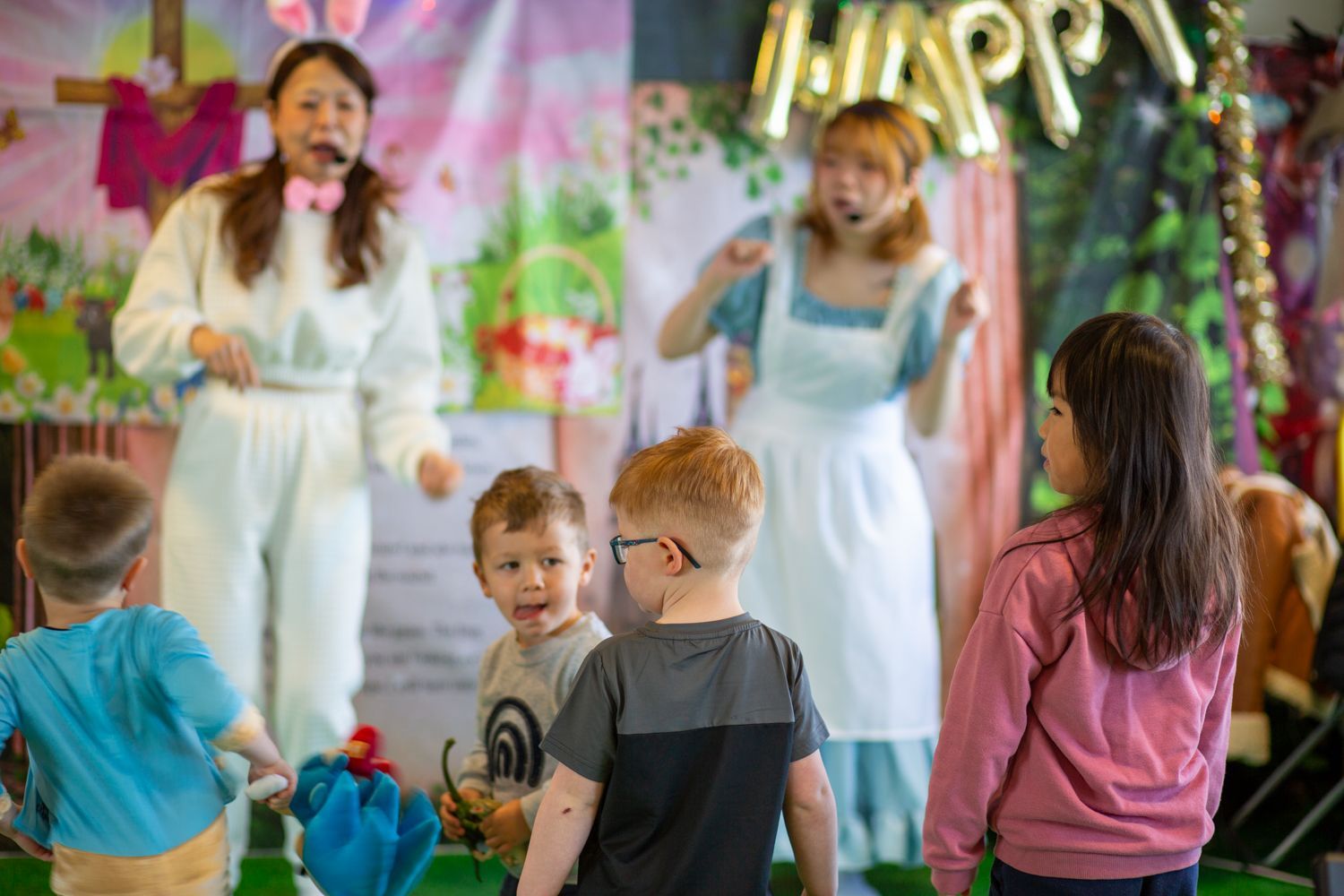Children watch two women in costumes on a stage. One wears a bunny suit, the other an Alice in Wonderland dress.