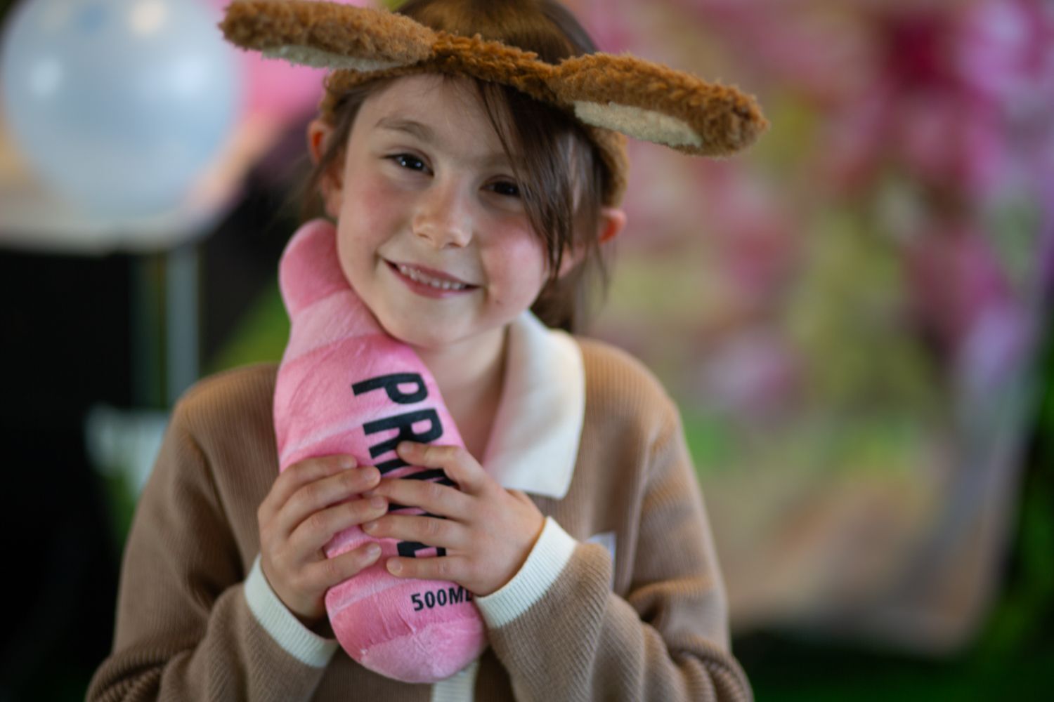 Girl in deer antlers, smiling, holding pink cast with “PRE” on it. Wearing brown sweater, blurred background.
