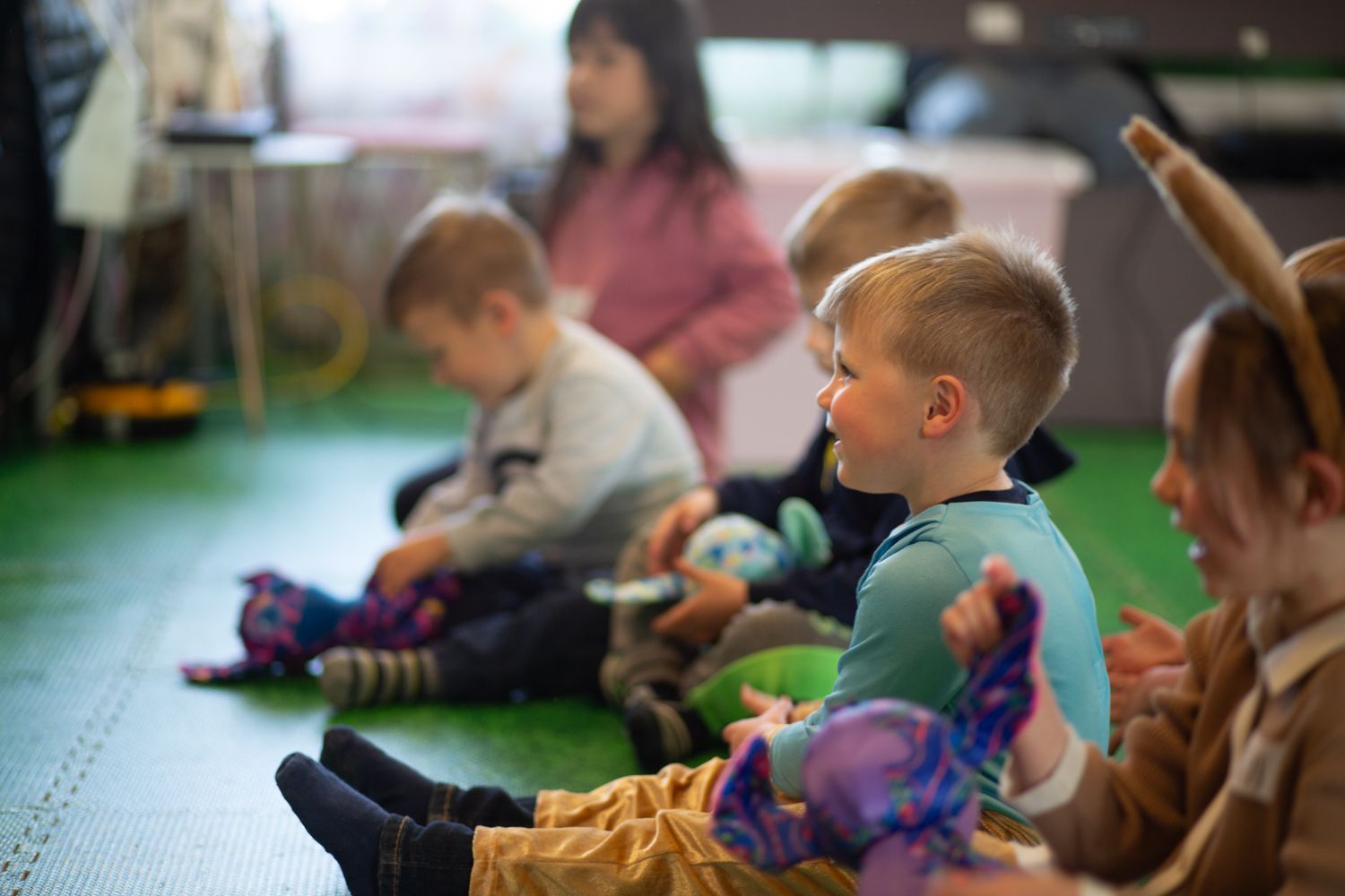 Children sit on a green floor, smiling, some holding toys. One wears bunny ears.