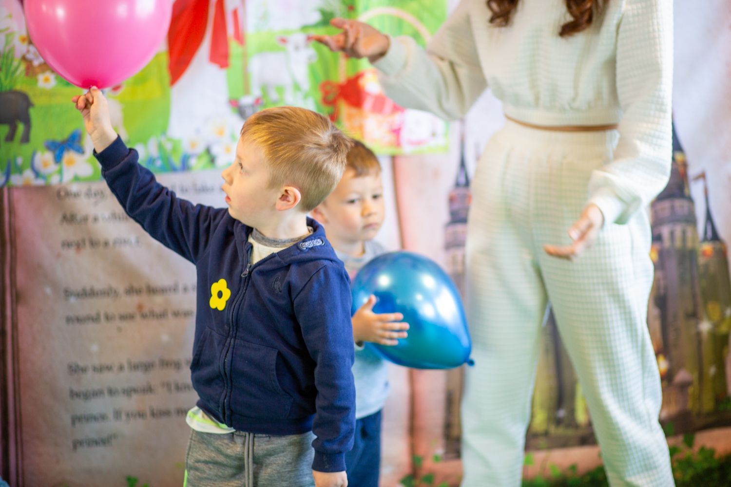 Boy raises pink balloon. Another child holds blue balloon, near a woman. Party setting.