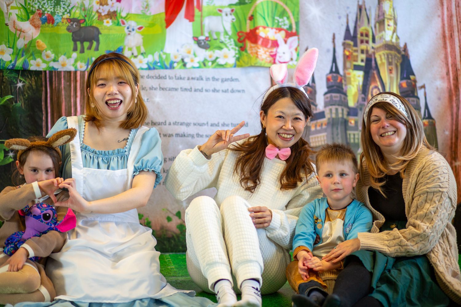 Group of adults and children in costumes posing, smiling, and making hand gestures in front of a fairytale backdrop.