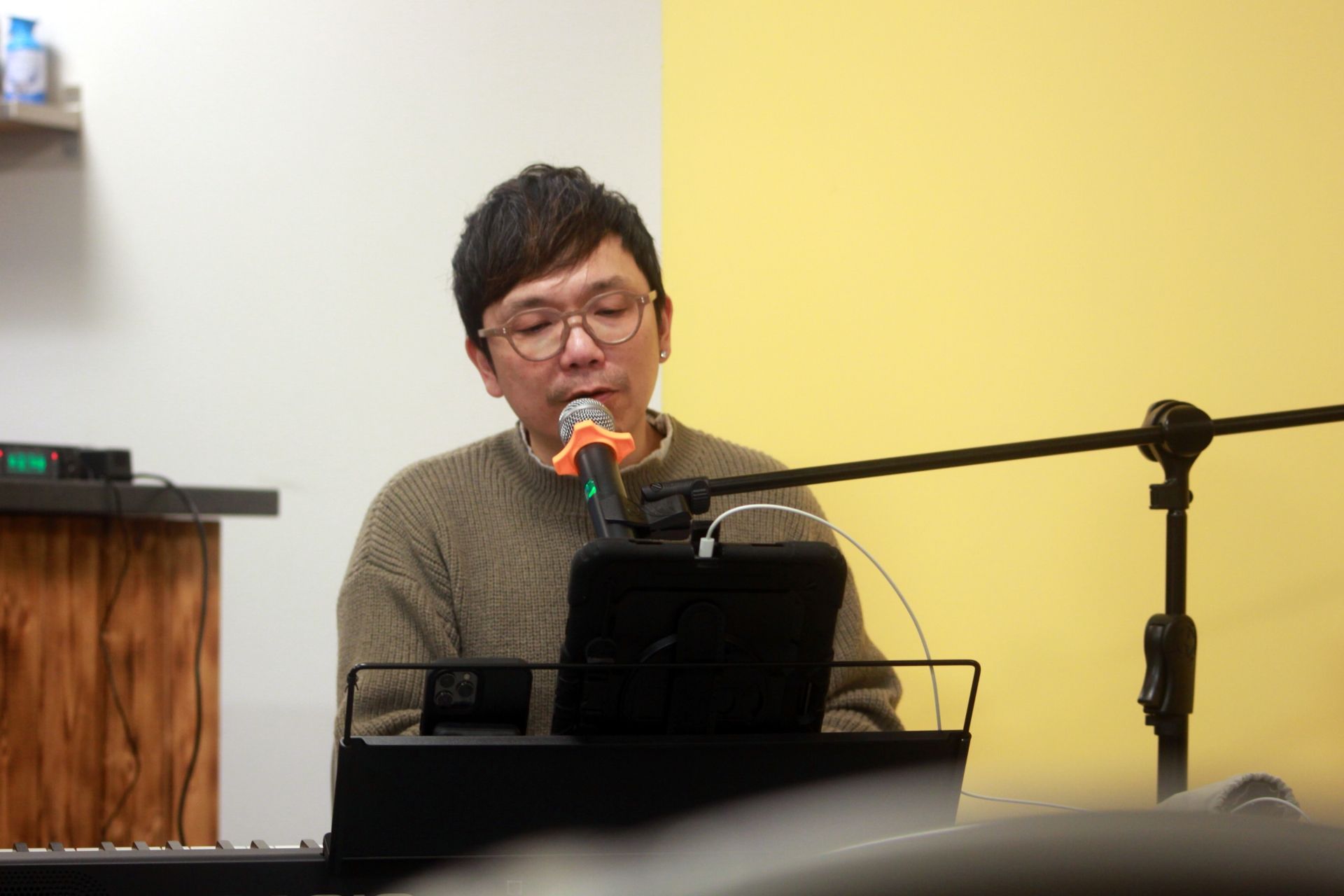 Man with glasses sings into a microphone while playing a keyboard; yellow and cream background.