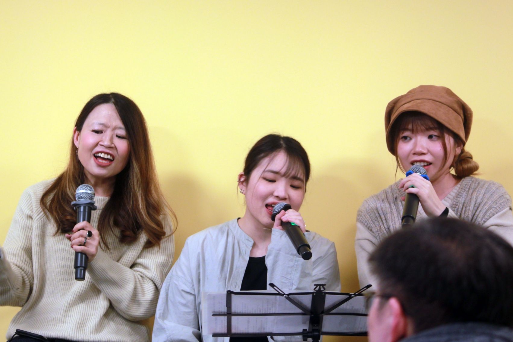 Three Asian women singing into microphones, standing in front of a yellow wall, while performing on stage.
