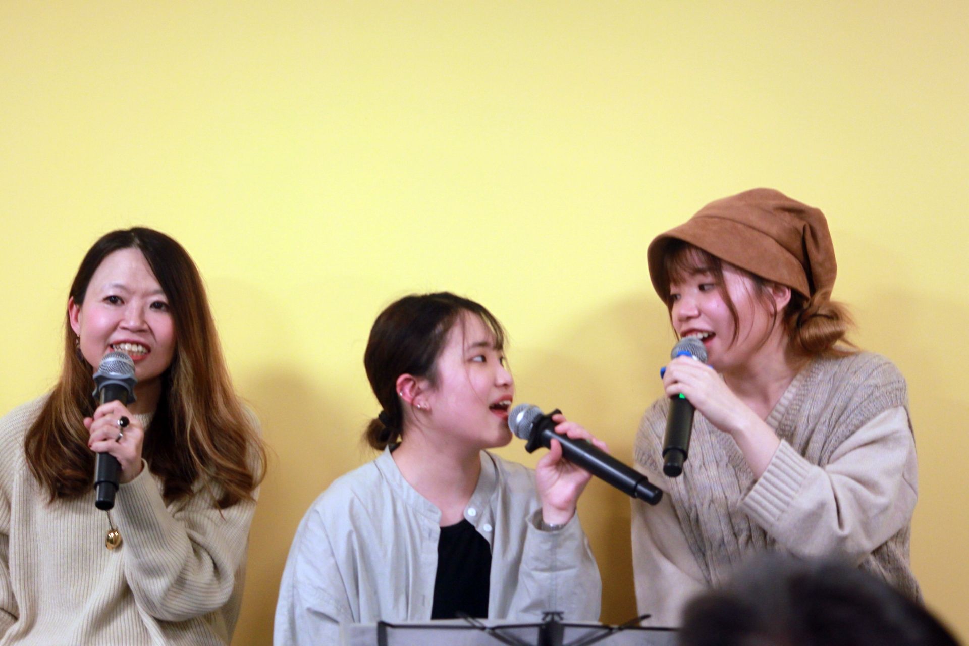 Three women singing into microphones against a yellow background. They appear to be performing.