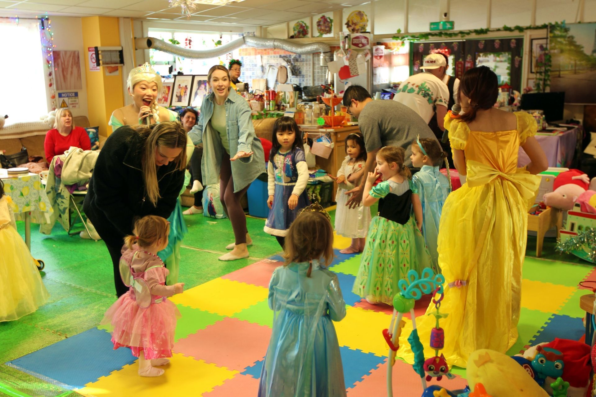 Children in princess costumes play at a party, adults supervise in a colorful room.