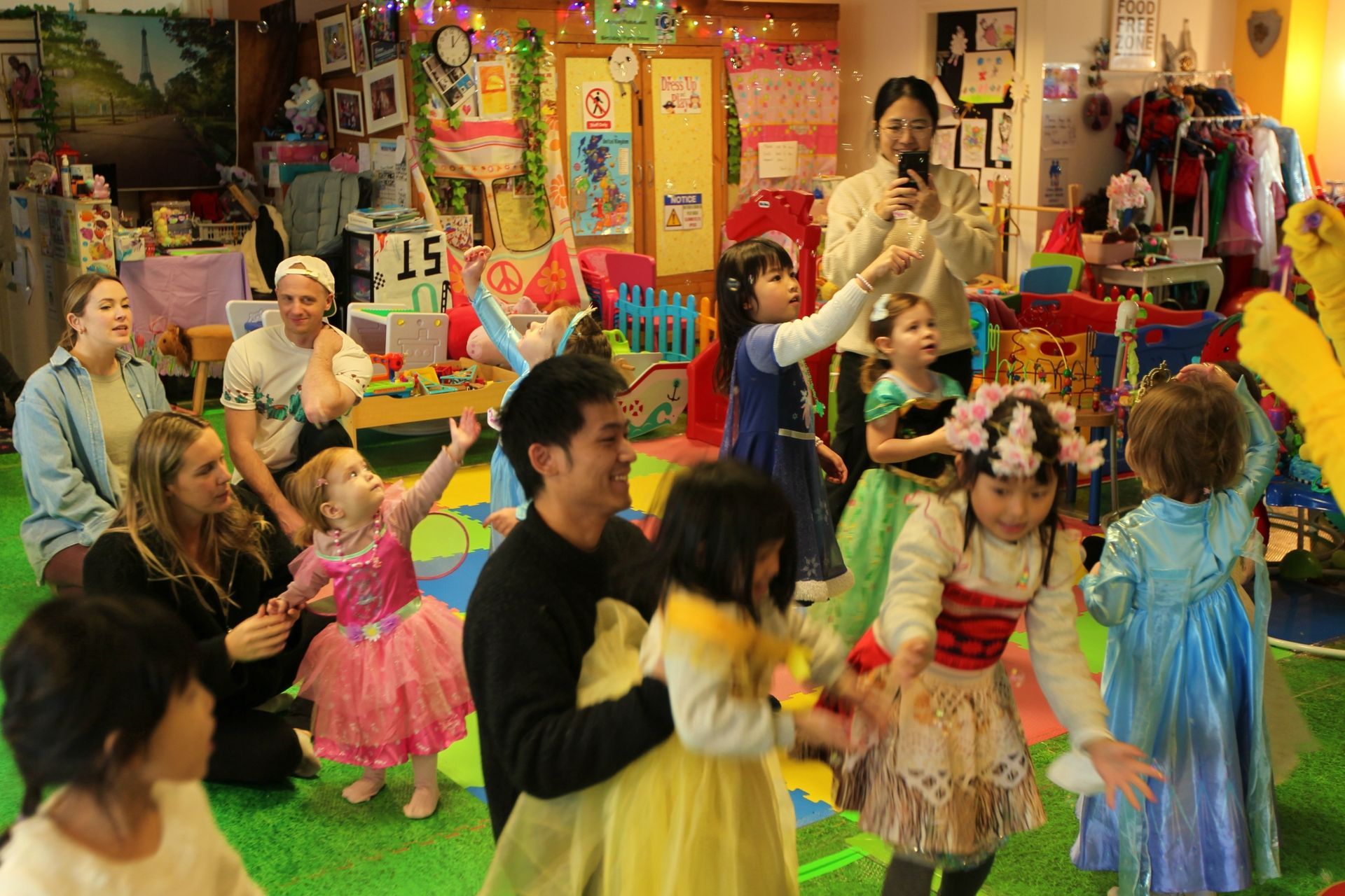 Children and adults in costumes, celebrating in a colorful indoor playroom.