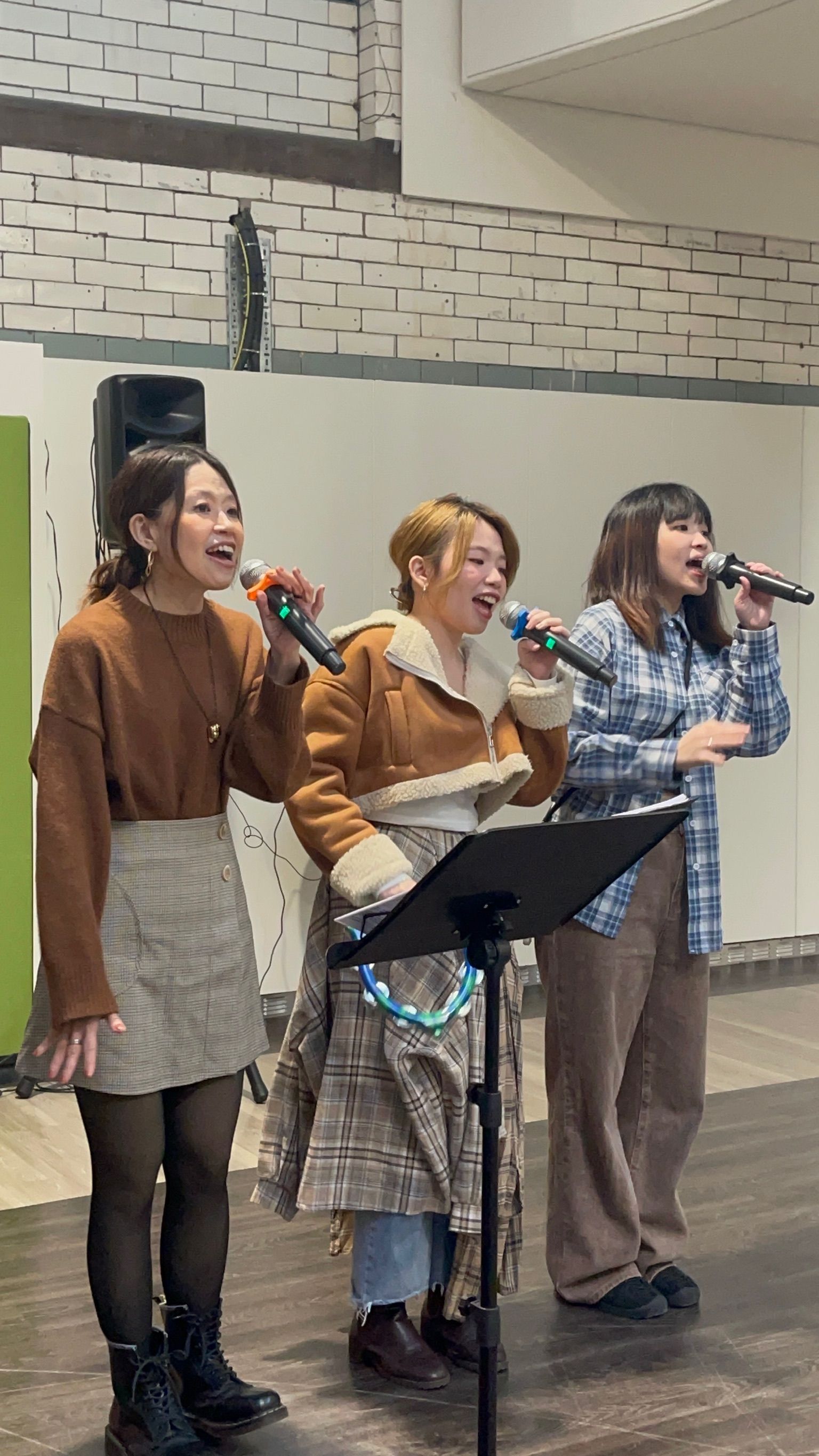 Three women singing into microphones, standing on a stage. They wear casual clothes, are mid-song, and appear excited.