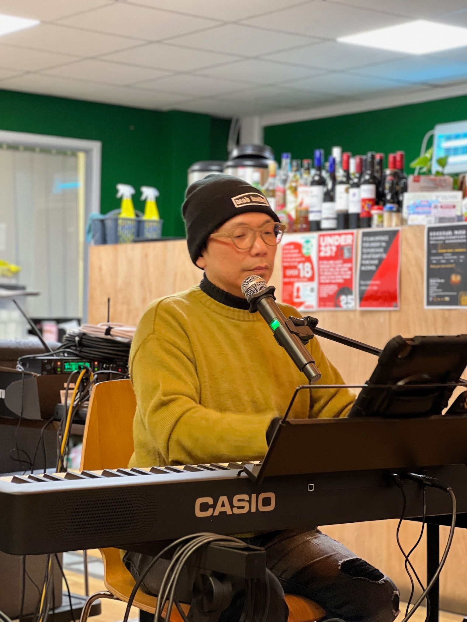 Man in beanie plays a Casio keyboard, singing into a microphone in a cafe setting.