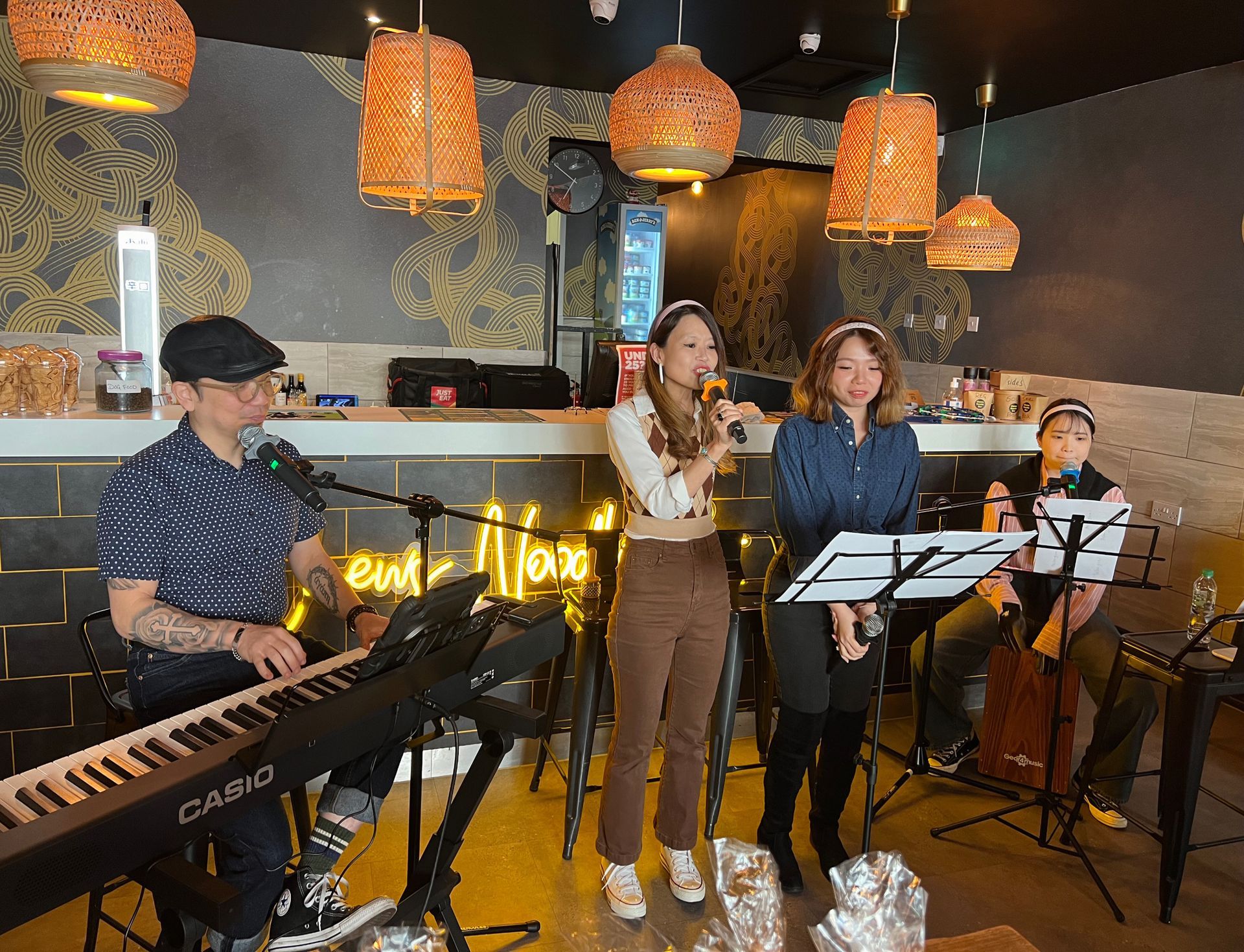 A band performs inside a bar. A woman sings into a mic. Other band members play keyboard and percussion.