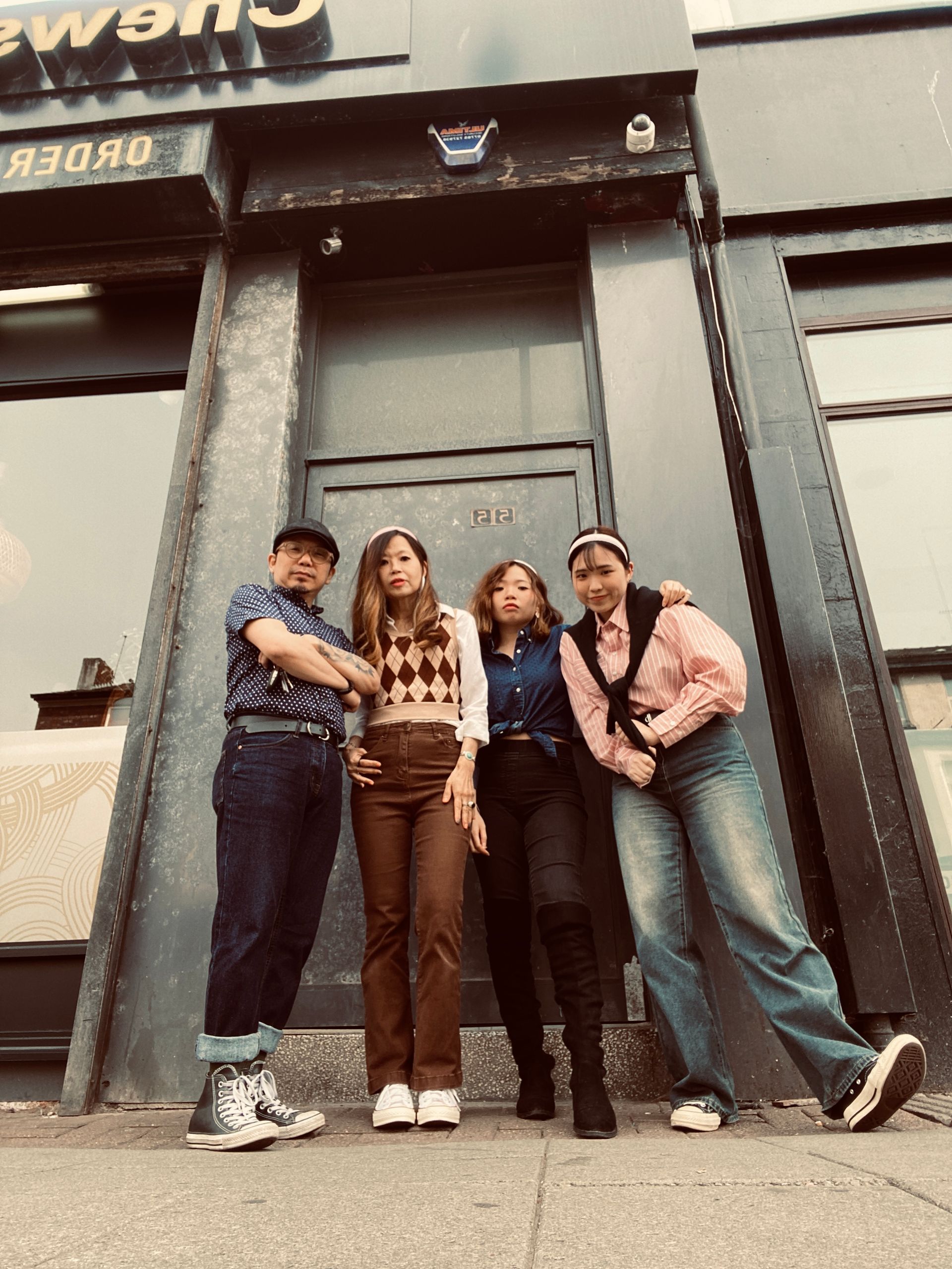 Four people in vintage outfits pose outside a storefront; sunny day, neutral tones.