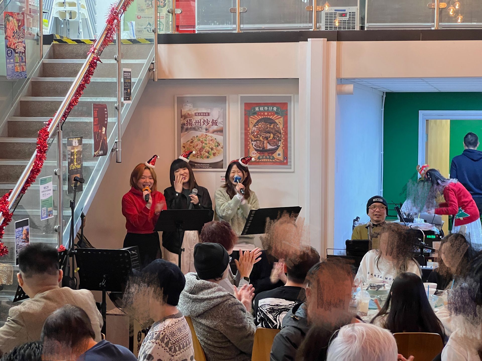 A band of three women sing onstage with a crowd watching, Christmas decorations in a venue.
