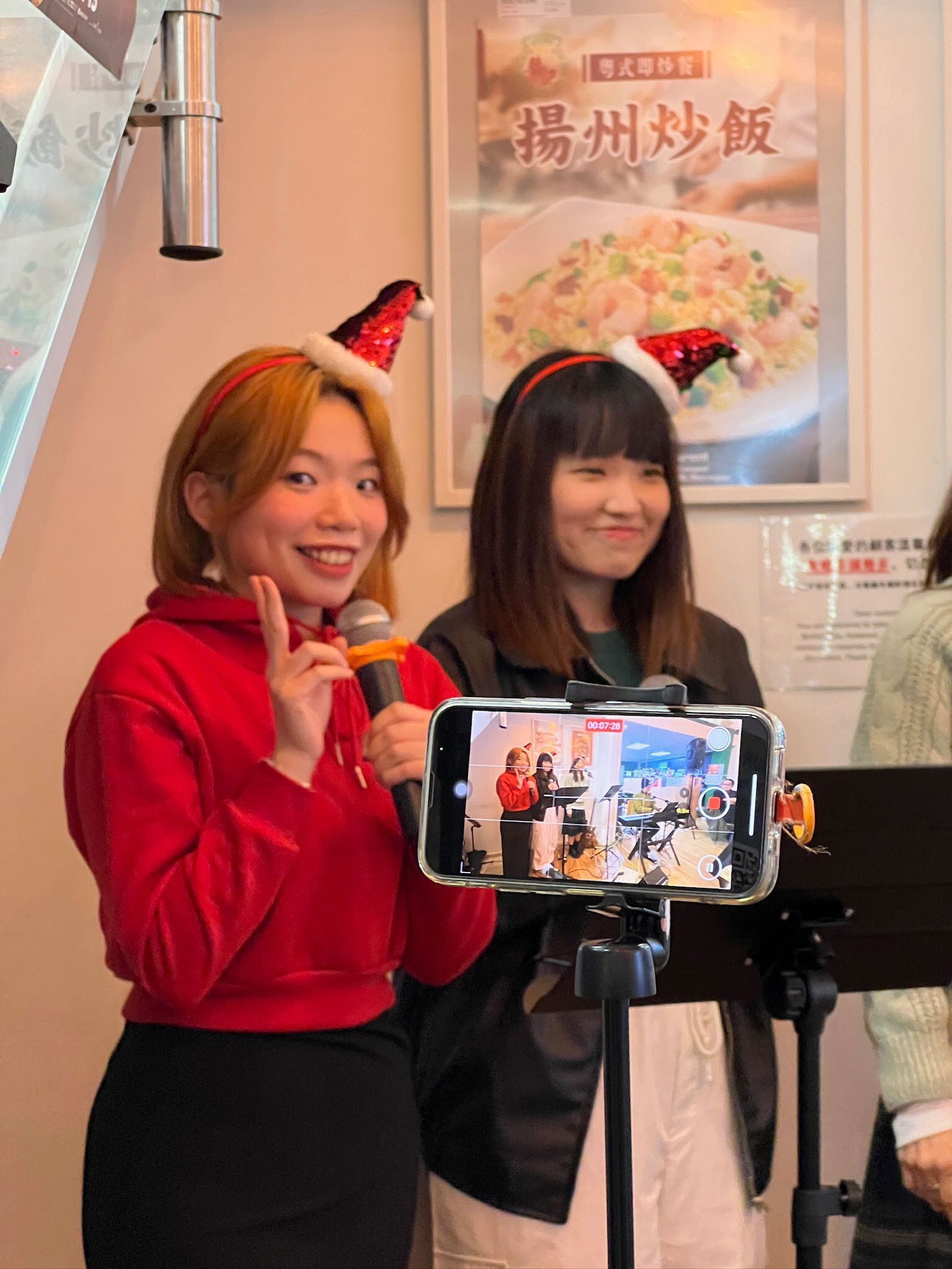 Two women in Santa hats live streaming. One holds a microphone, the other holds a phone on a tripod.