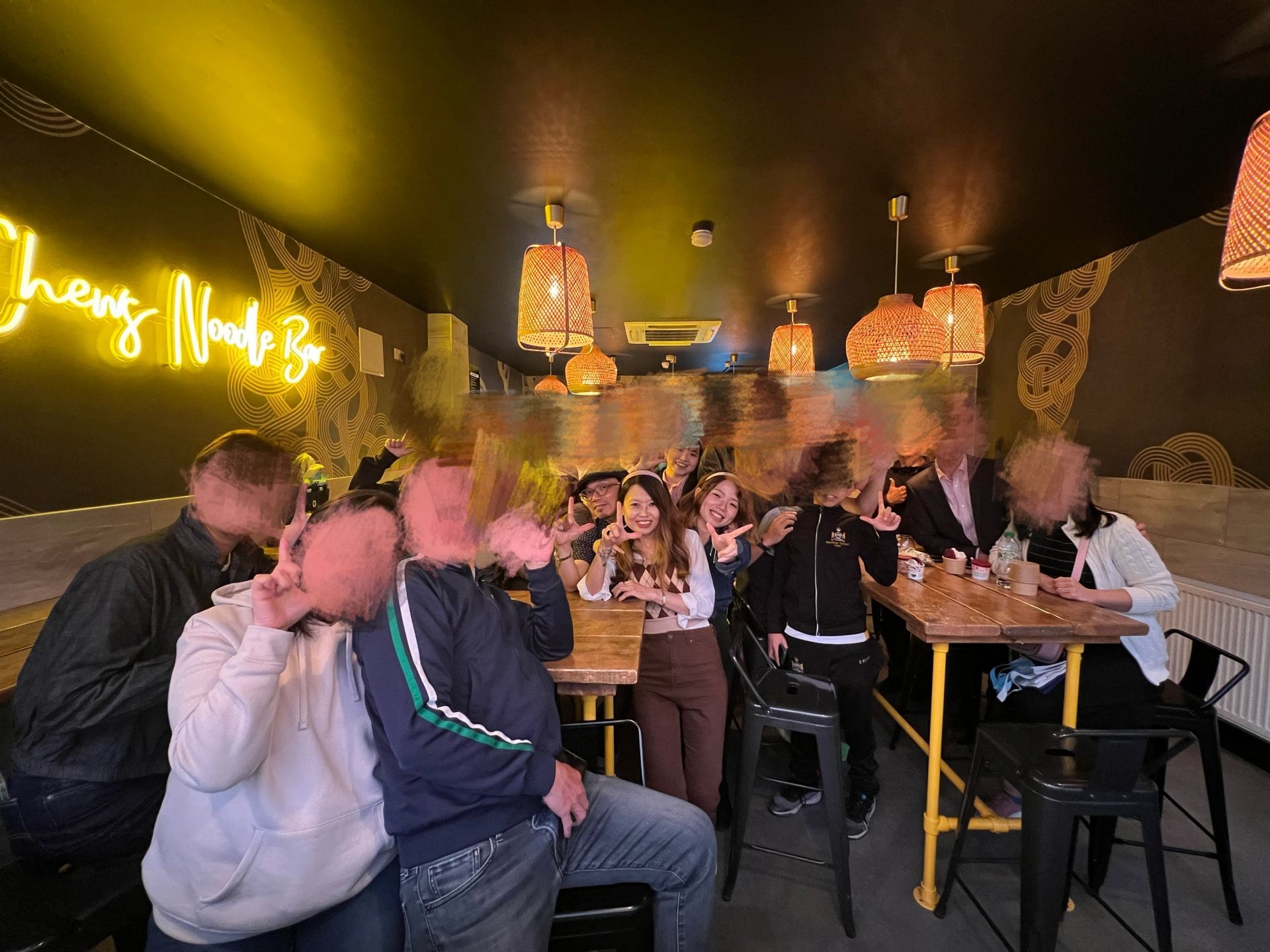 People at a restaurant, one woman smiling at the camera, others partially blurred. Yellow neon sign and hanging lights.