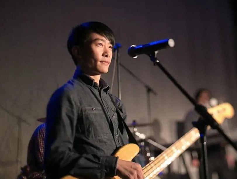 Man playing bass guitar on stage, looking upward. He's wearing a blue denim shirt.