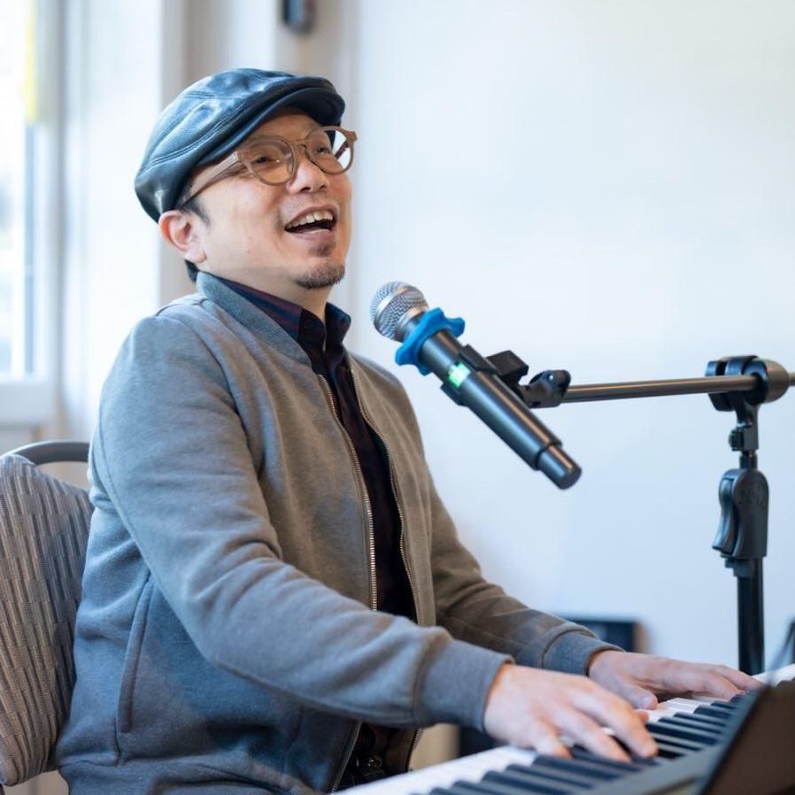 Man playing piano and singing into a microphone, wearing glasses, hat, and gray jacket.