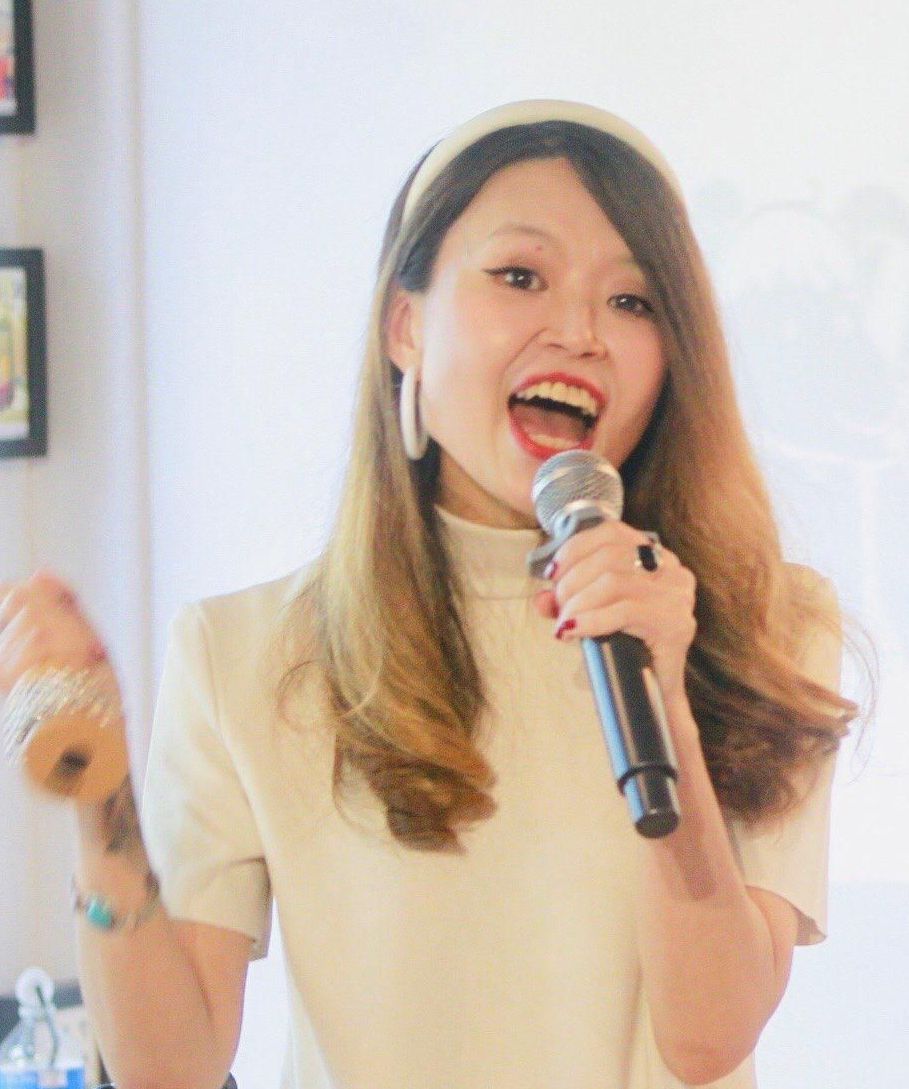 Woman singing into a microphone, wearing a white outfit and headband, bright setting.