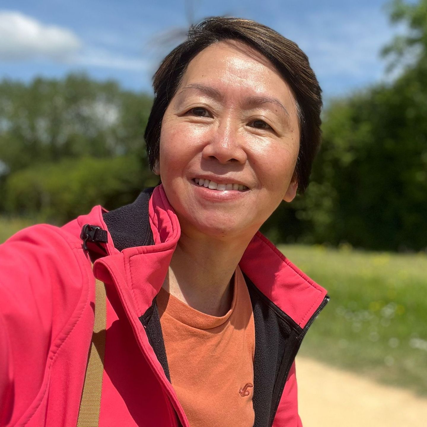 Woman with short brown hair smiles, wearing a red jacket and orange shirt outdoors on a sunny day.