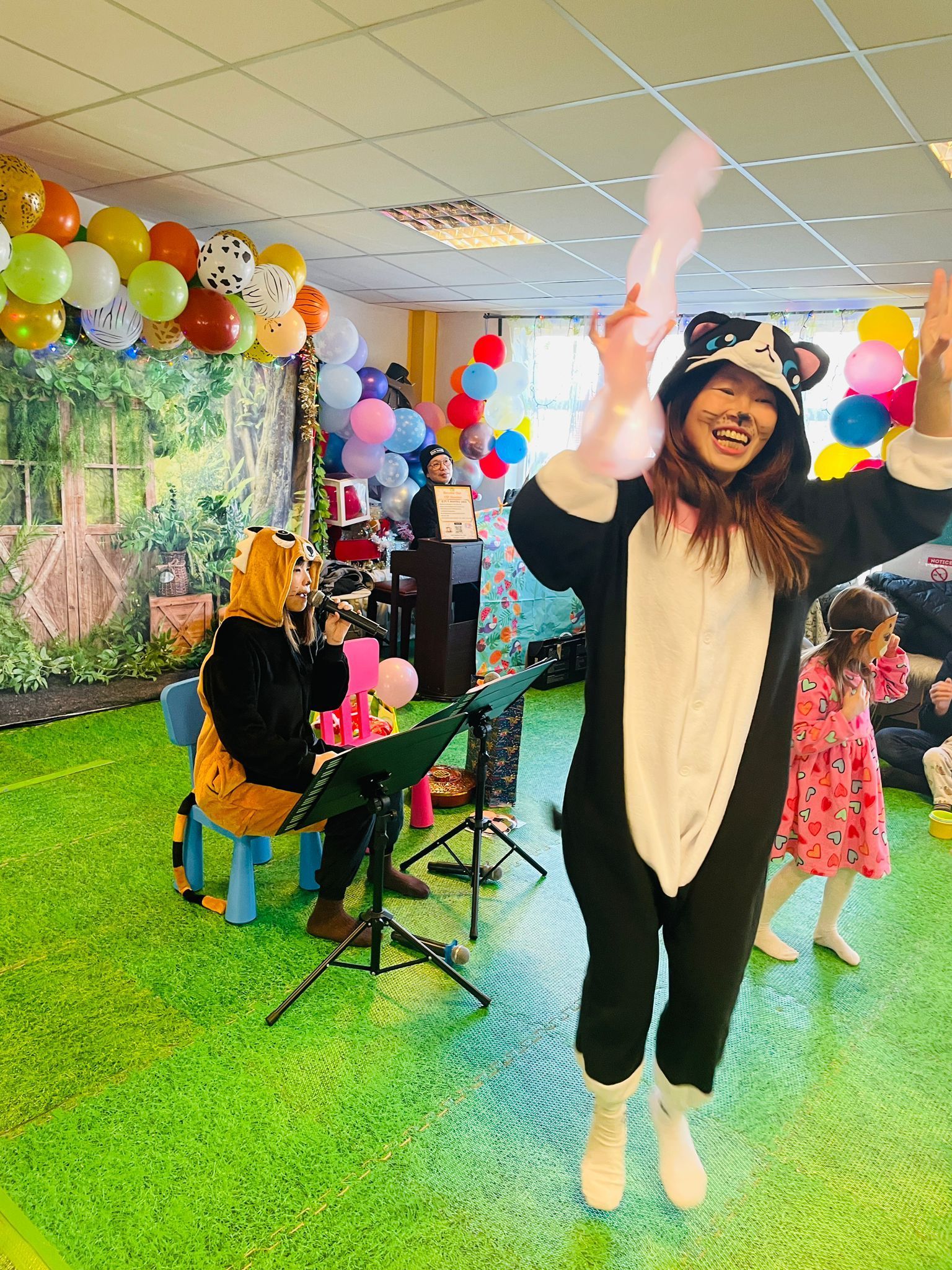 Person in cow costume celebrates at a party; musician at piano; colorful balloons.