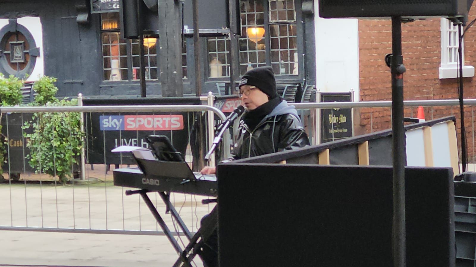 A man plays a keyboard and sings outdoors, wearing a black jacket and hat, in front of a building with signs.