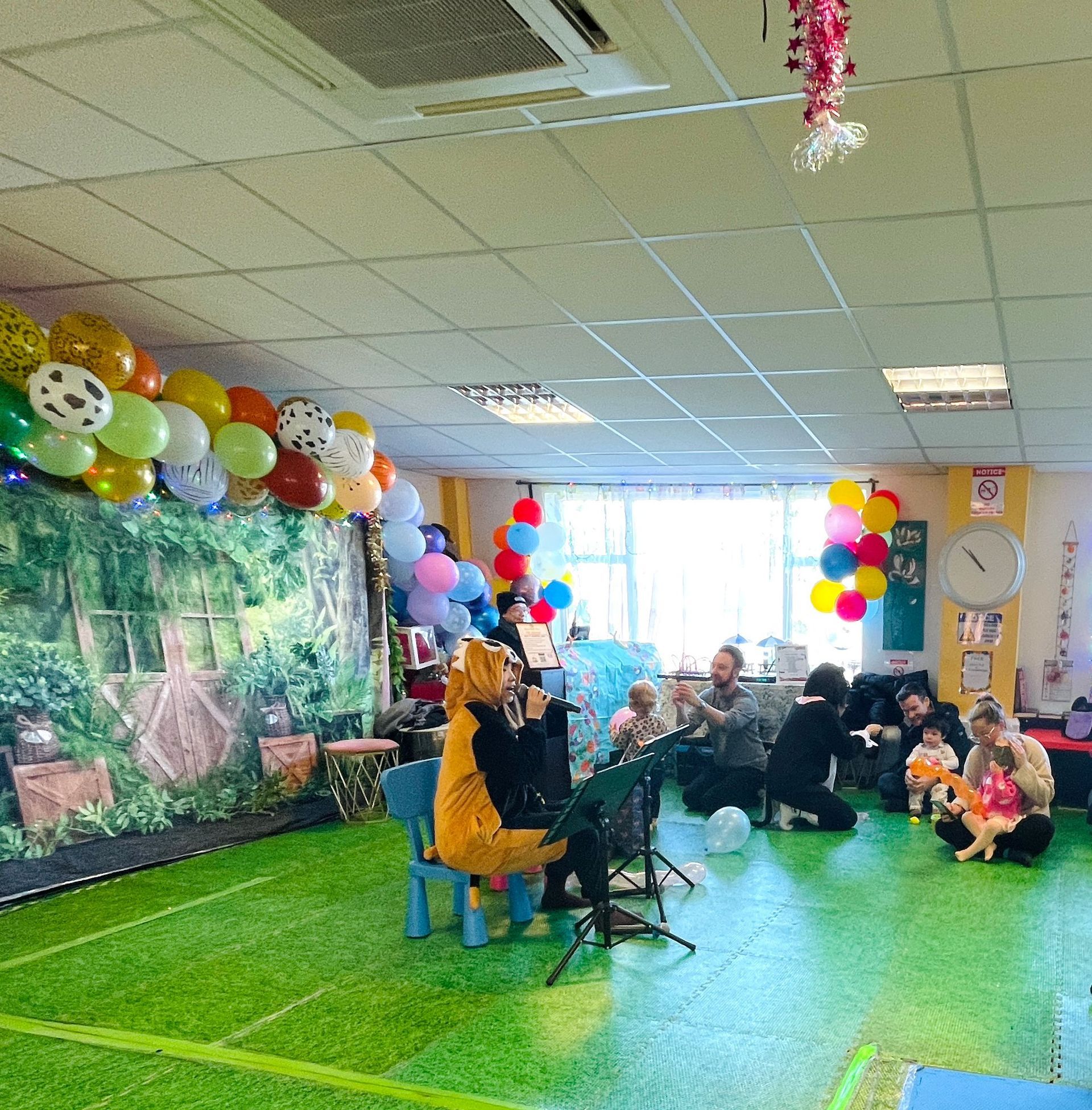 Children's party: musician in yellow costume plays for seated kids, room decorated with balloons and artificial grass.