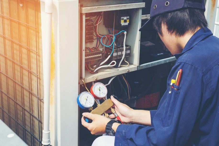 HVAC technician in blue uniform servicing outdoor AC unit, using gauges.