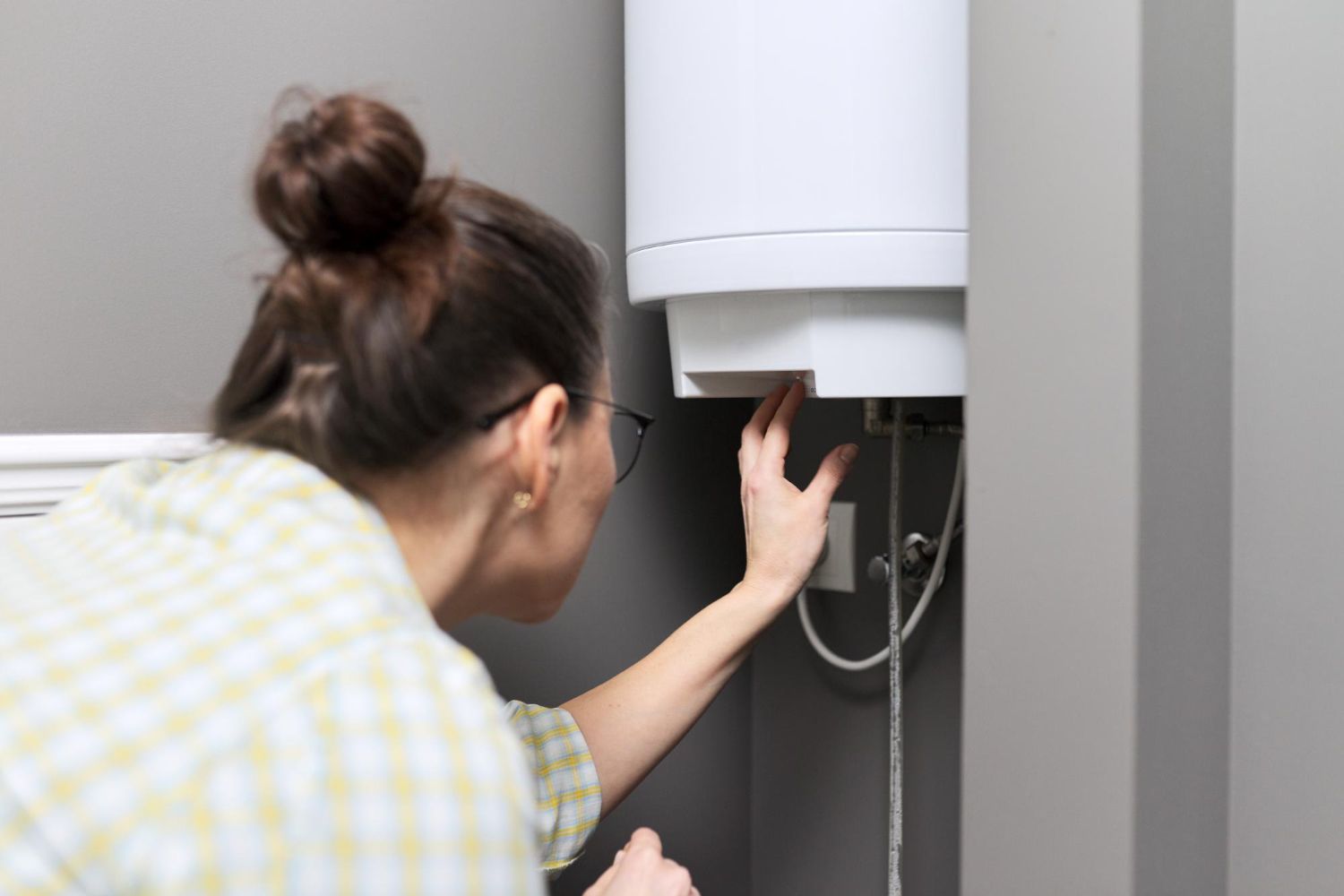 Woman inspecting a white water heater in a gray room. She wears glasses and has a bun, hand reaching toward the appliance.