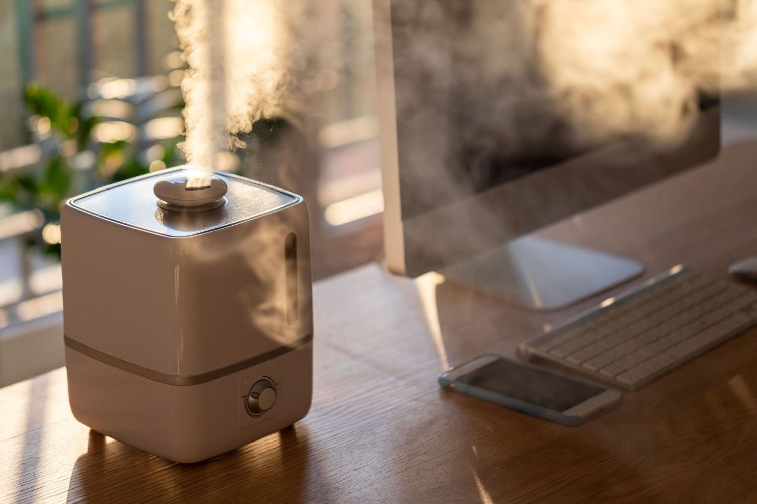 White humidifier emitting steam on a wooden desk near a computer and phone.