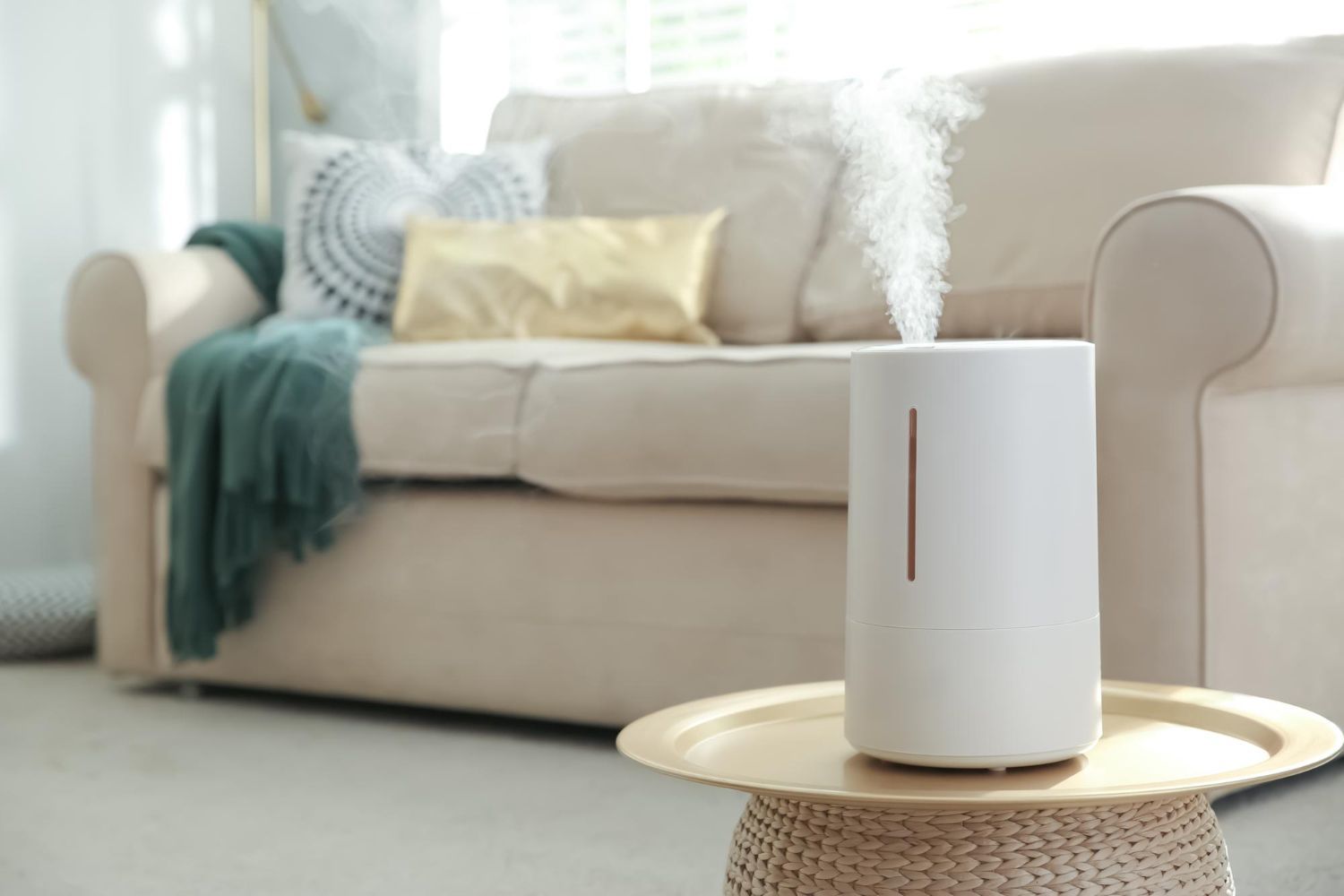 Humidifier emitting steam on a side table in front of a beige couch with pillows and a blanket.
