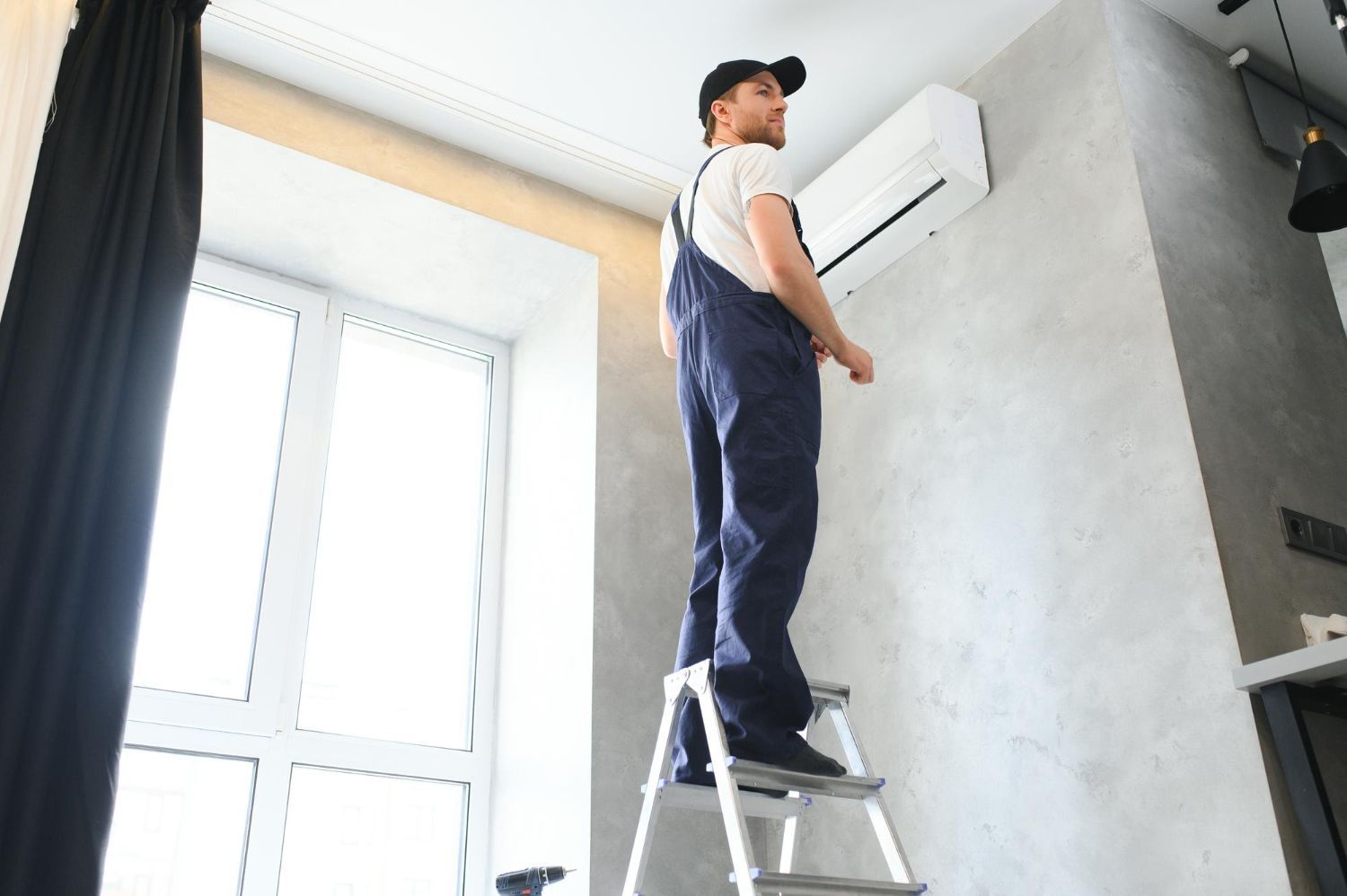 Person in blue overalls on a ladder inspecting an air conditioner unit mounted on a wall.