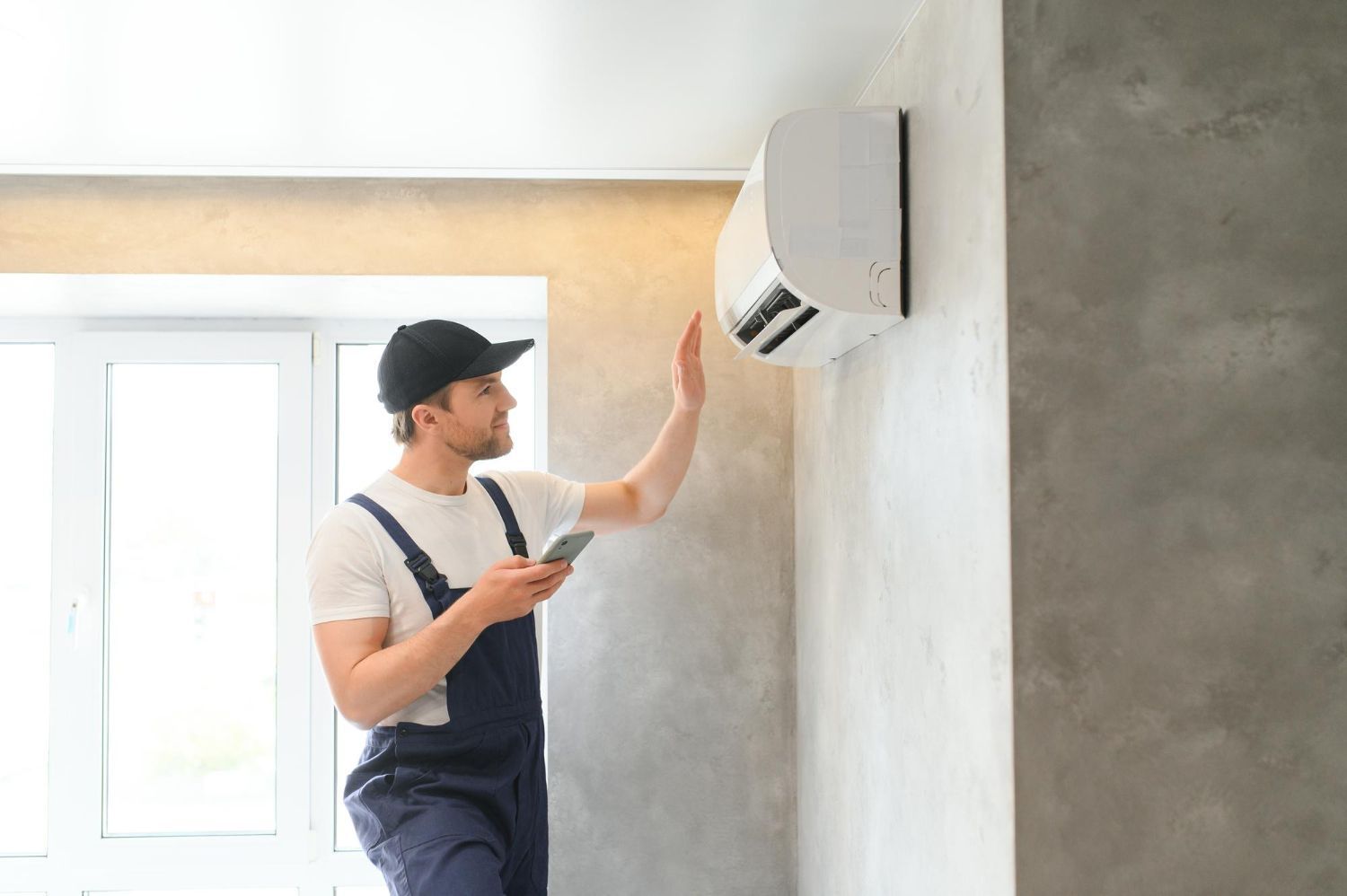 HVAC technician in overalls checks a wall-mounted air conditioner with a phone, reaching out to it, indoors.