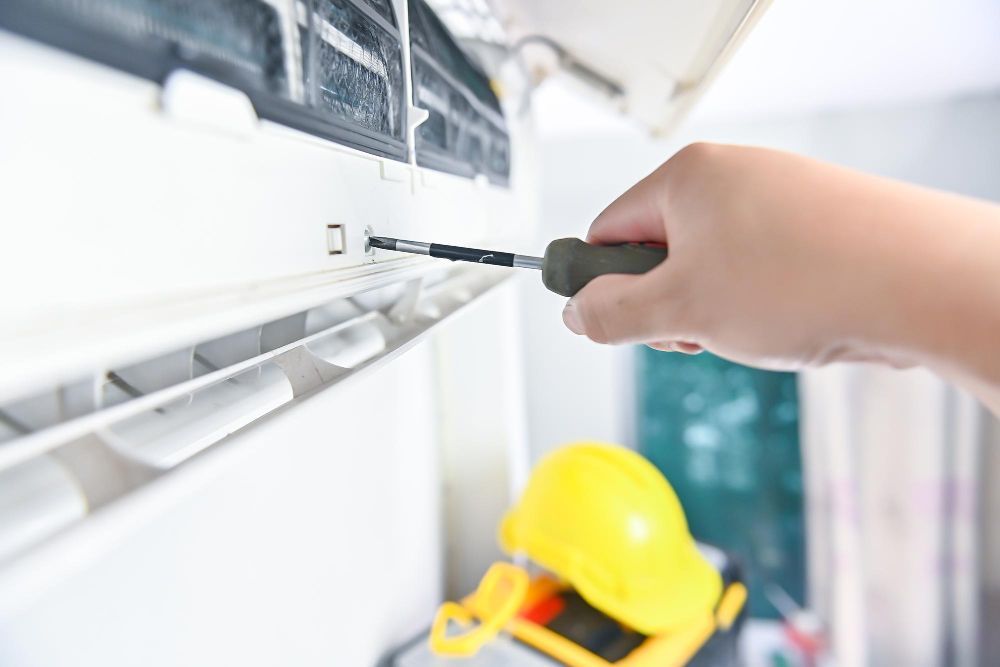 Hand using screwdriver to fix white air conditioner. Yellow hard hat on shelf below.