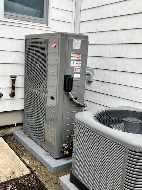 Two air conditioning units, beige, beside a building on a concrete pad with grass.