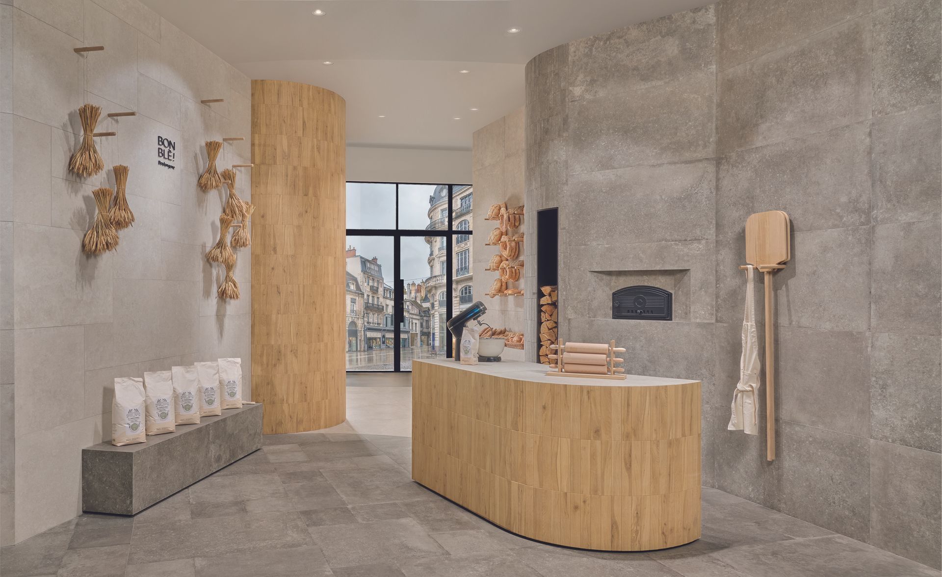 Interieur van de bakkerij: toonbank en pilaar van licht hout, grijze muren, brood tentoongesteld.