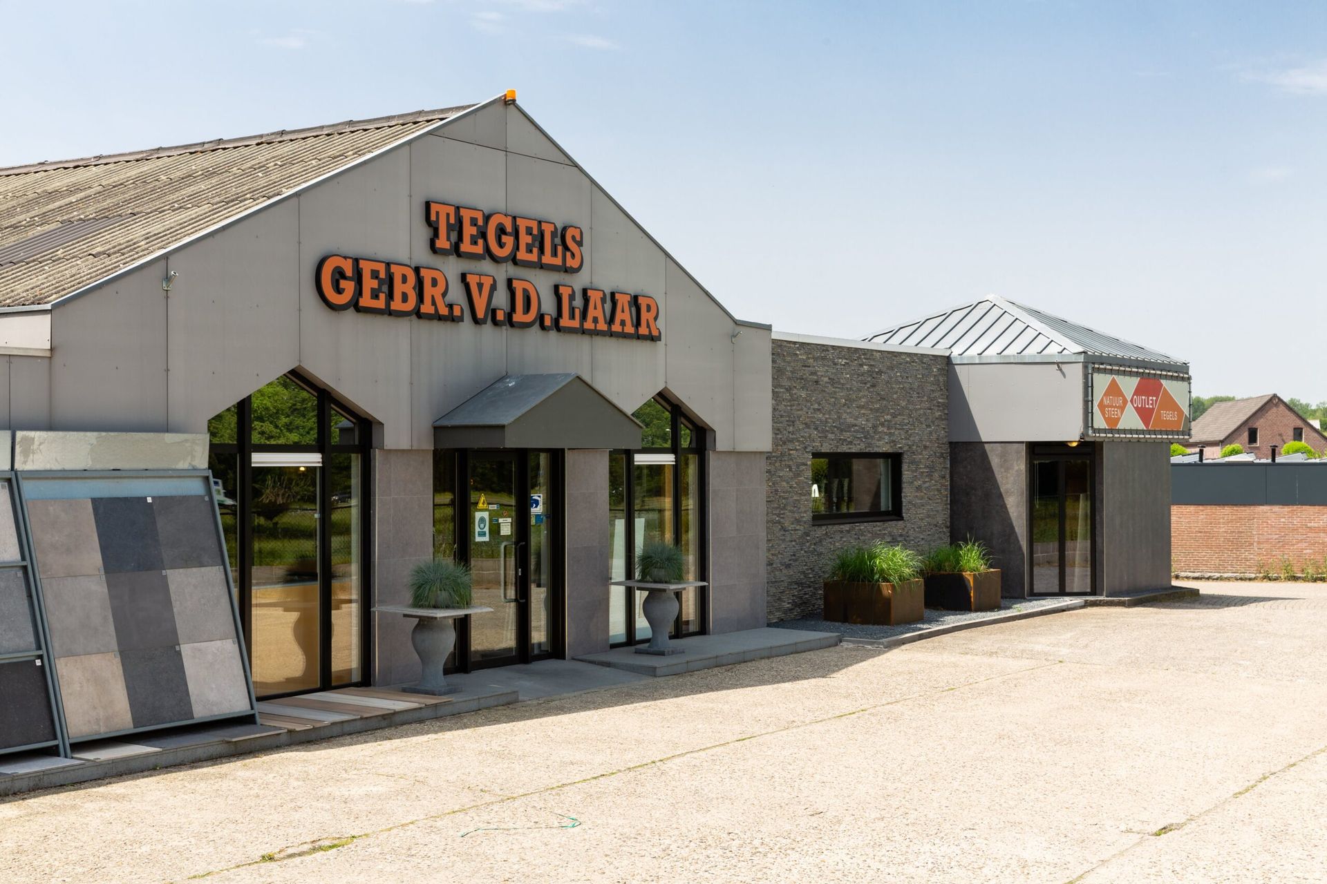 De buitenkant van een tegelwinkel, "TEGELS GEBR. VD HAA." Grijs gebouw met glazen deuren en een etalage met tegels.