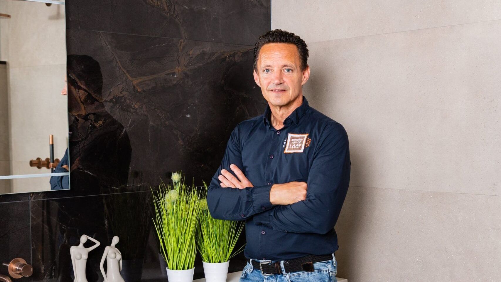 Een man in een blauw shirt met gekruiste armen staat in een badkamer.