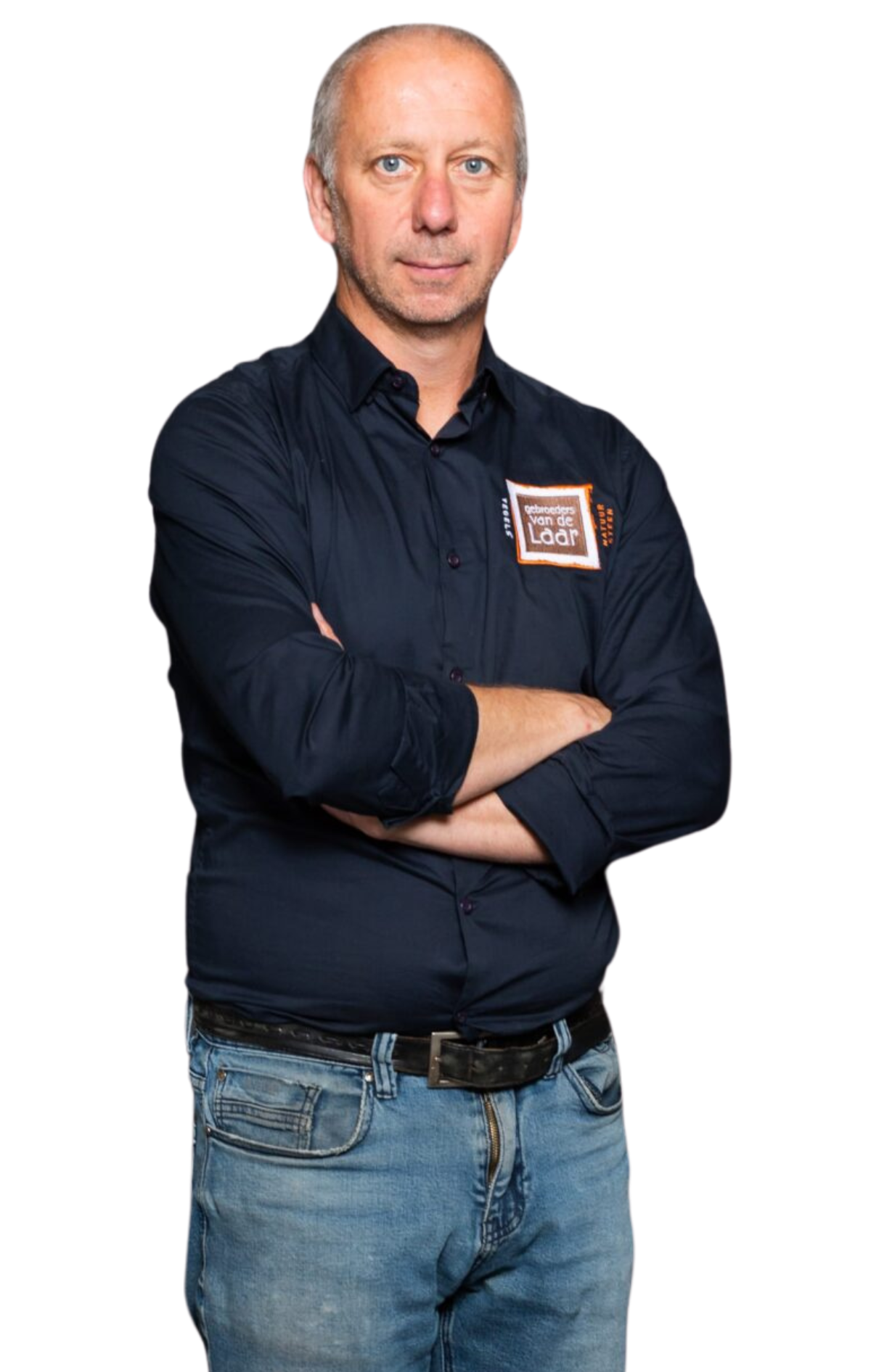 Een man met gekruiste armen, gekleed in een donkerblauw shirt en spijkerbroek, staat tegen een witte achtergrond.