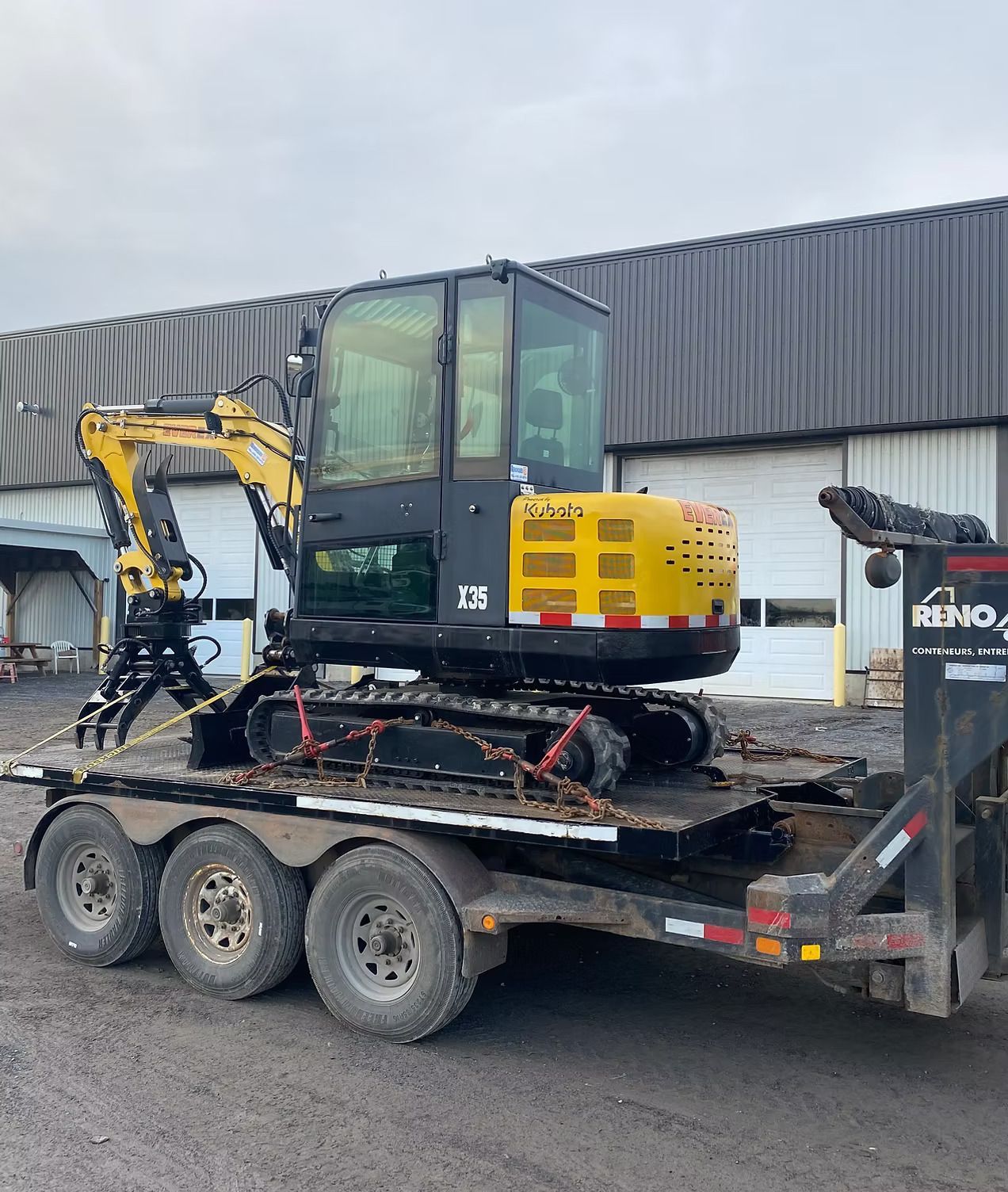 Une excavatrice jaune est garée sur une remorque devant un bâtiment.
