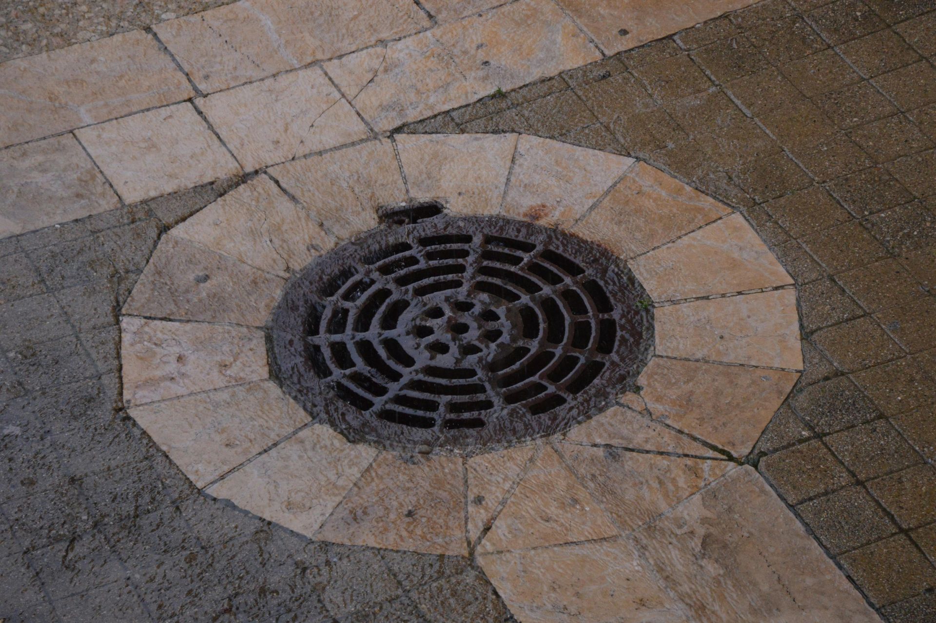 A circular metal drain cover surrounded by beige stone pavers on a textured sidewalk.