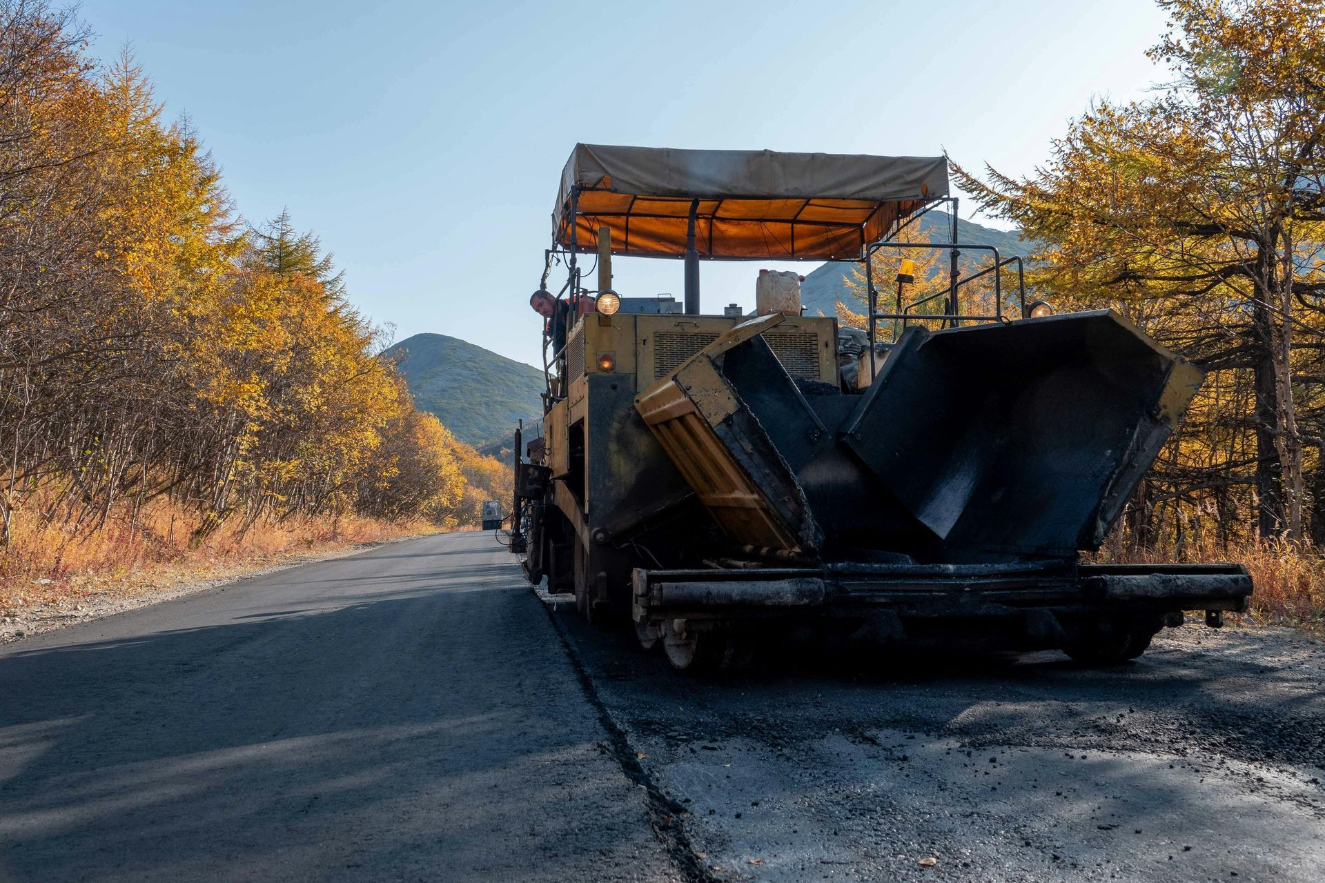 A yellow road paver sits on an asphalt road surrounded by autumn trees and mountains.