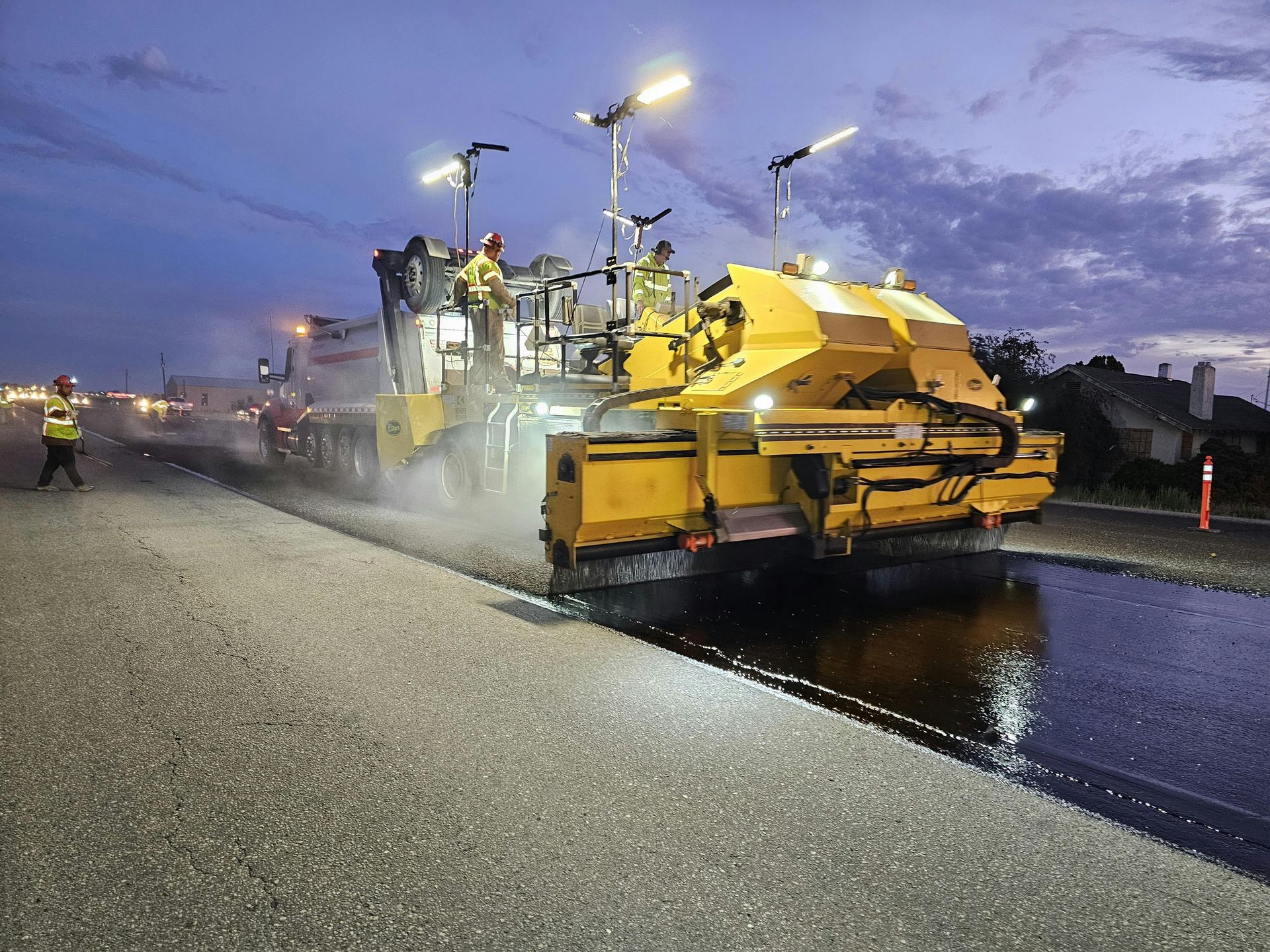 A yellow construction machine paves a road at twilight, with crew members nearby and wet asphalt visible on the ground.