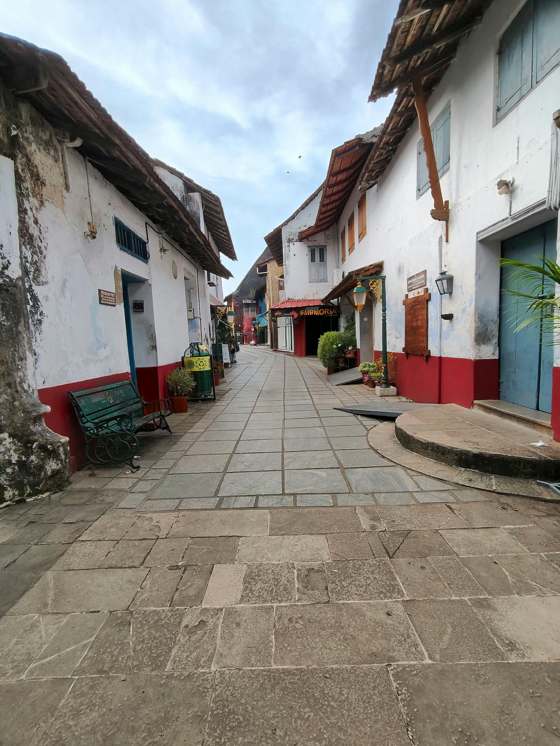 A narrow stone-paved street lined with white buildings featuring red trim, a green bench, and wooden roof overhangs.