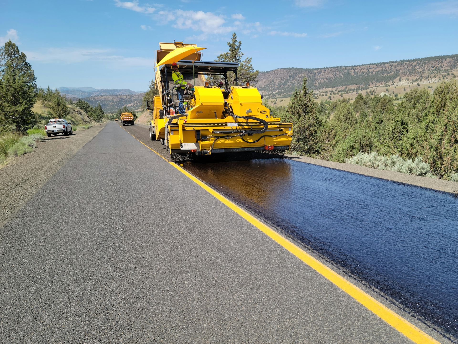 A bright yellow paving machine applies fresh asphalt to a road under a sunny sky near trees and hills.