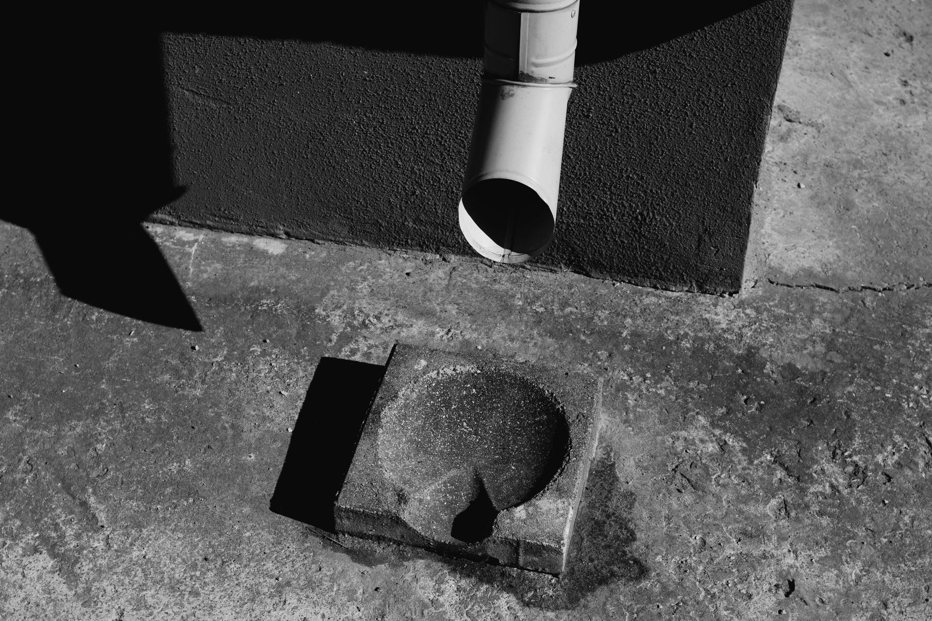 A black-and-white view of a downspout positioned above a small, square stone drainage basin on a concrete surface.