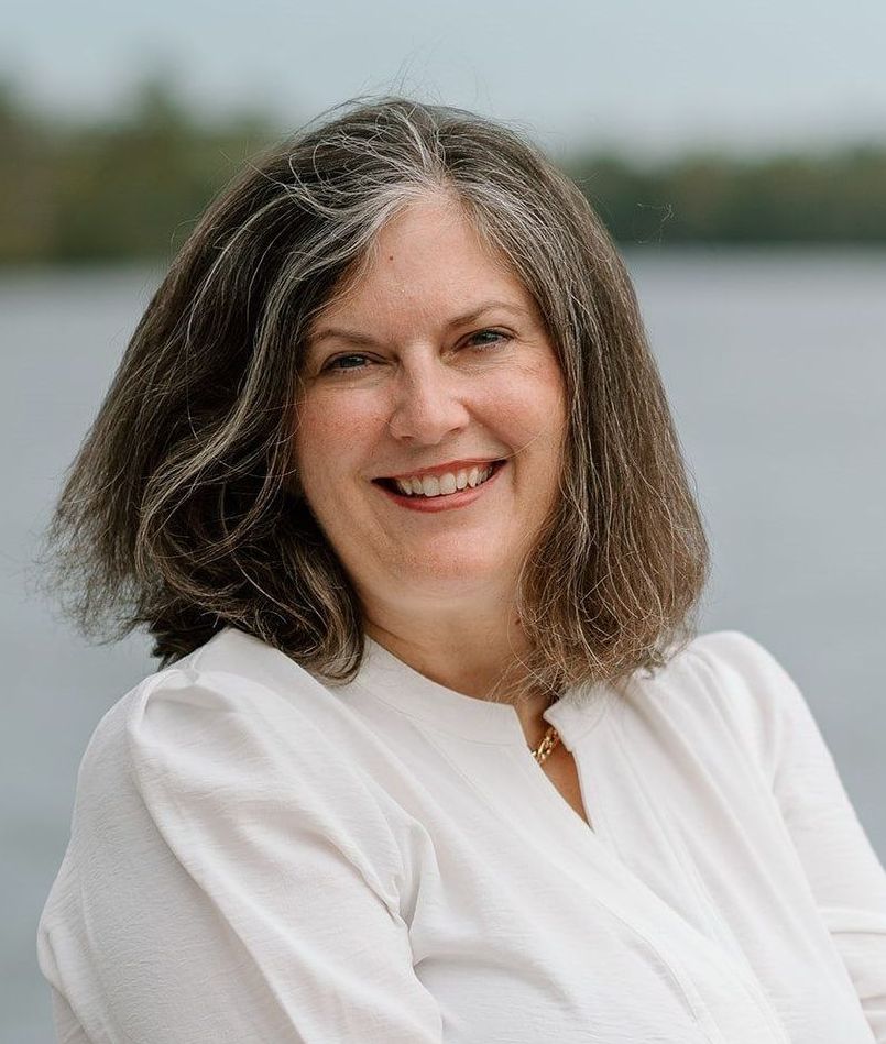 Lindsay, in a white shirt, is smiling in front of a body of water.