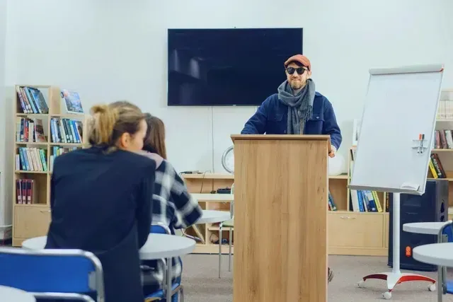 A man is standing at a podium in front of a group of people in a classroom.