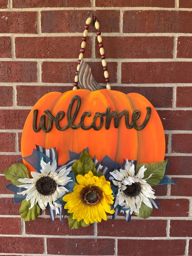 Orange pumpkin door sign with “welcome,” beaded hanger, and sunflowers against a brick wall