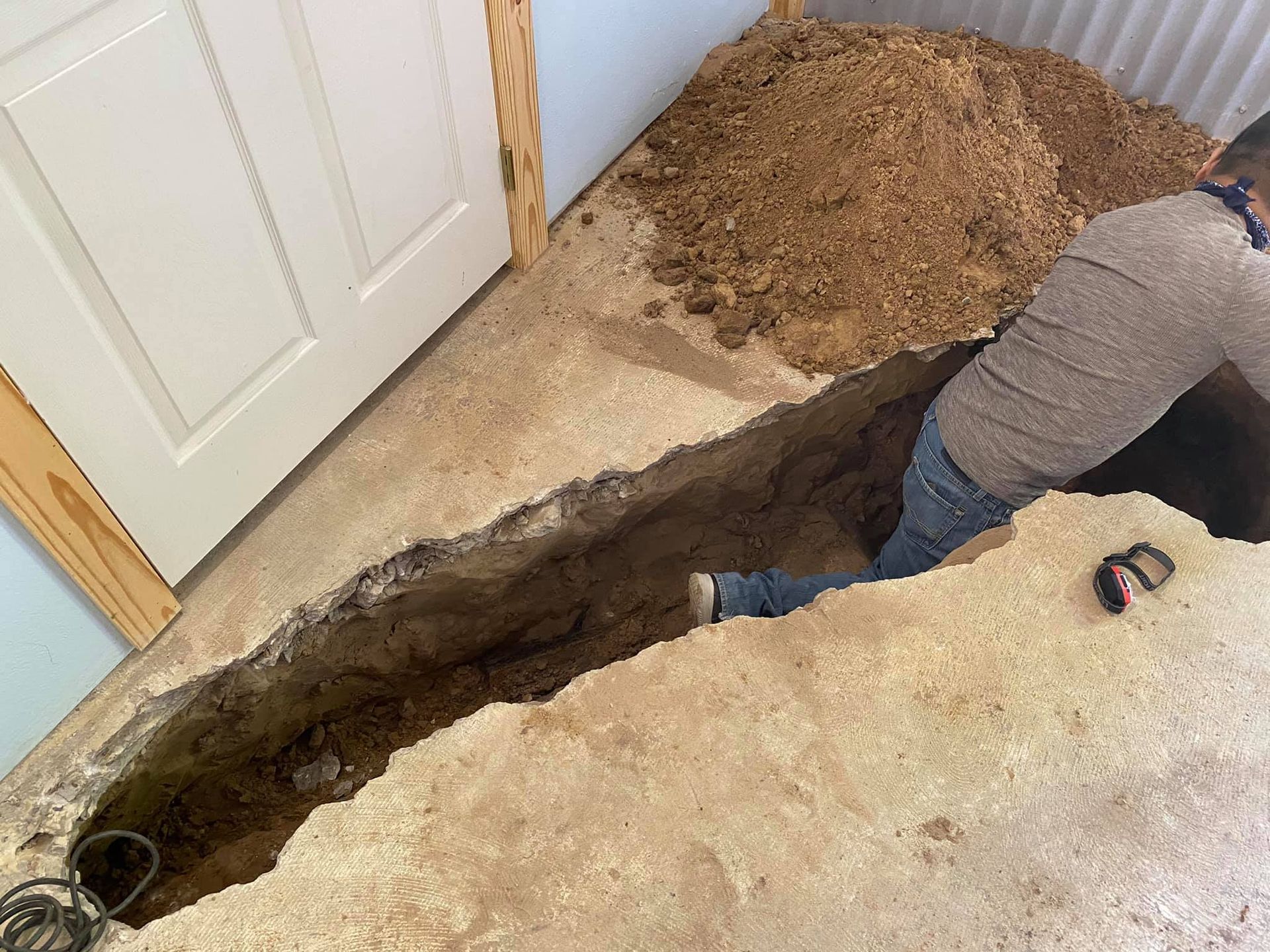 Person in a trench excavating near a doorway, surrounded by dirt and debris indoors.