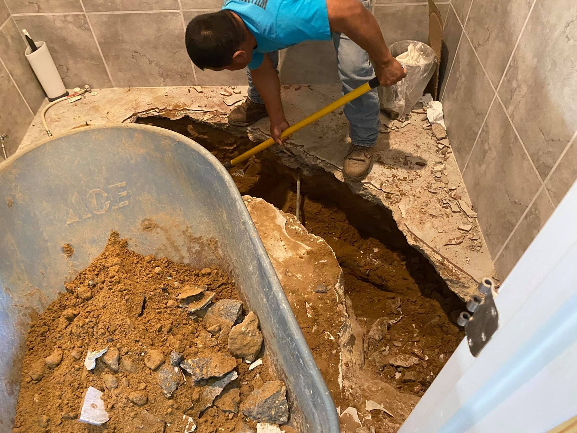 Person shoveling dirt from a trench in a bathroom floor into a wheelbarrow. Tile walls.