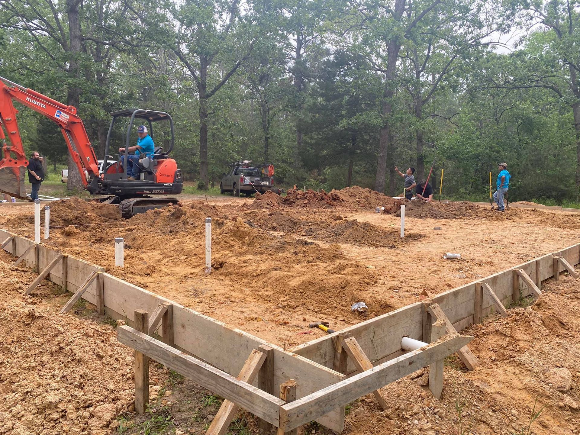 Construction site: foundation forms, excavator, workers, dirt, trees.